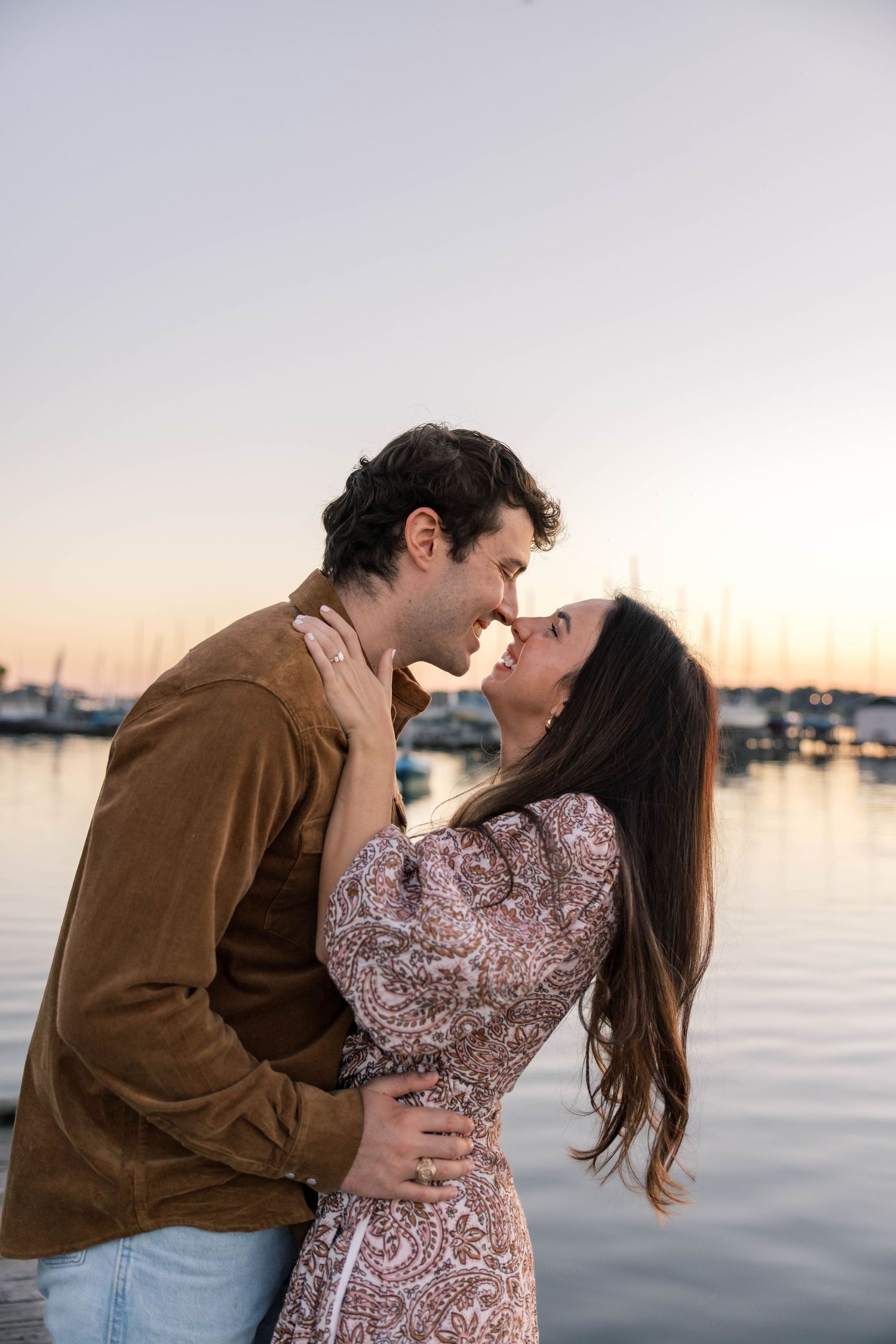 Couple sharing a tender almost-kiss embrace at golden hour by a marina with sailboats behind them.