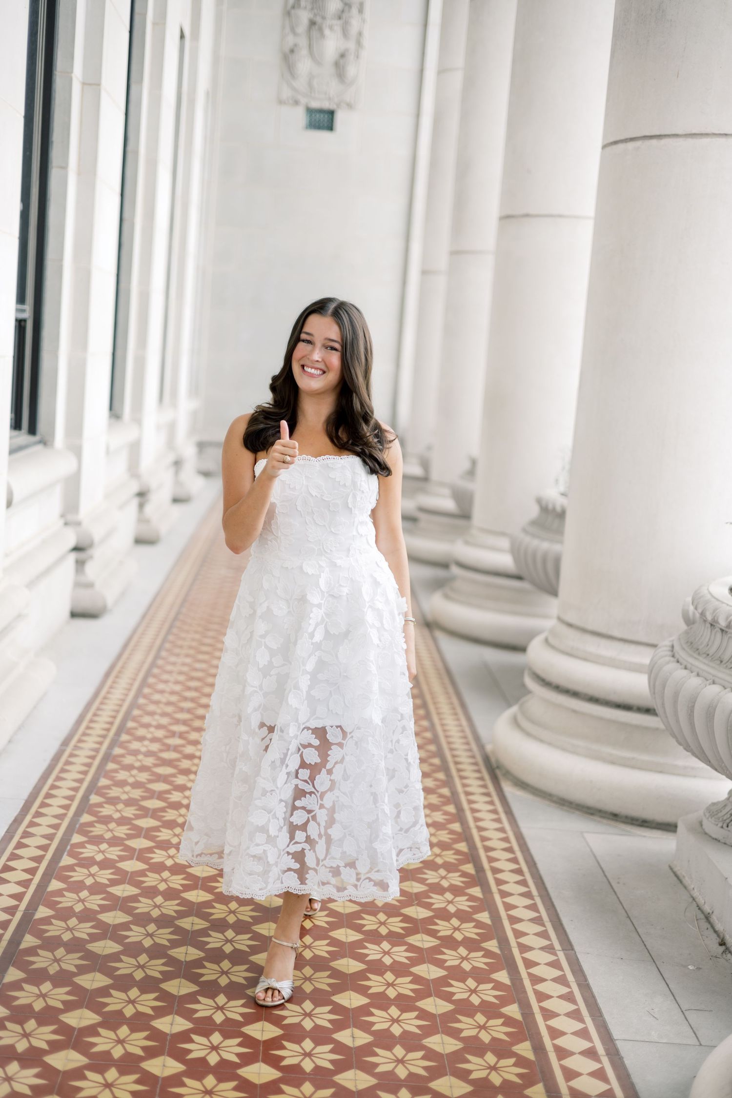 Cheerful graduate in white strapless lace dress gives thumbs up while walking in elegant columned corridor.