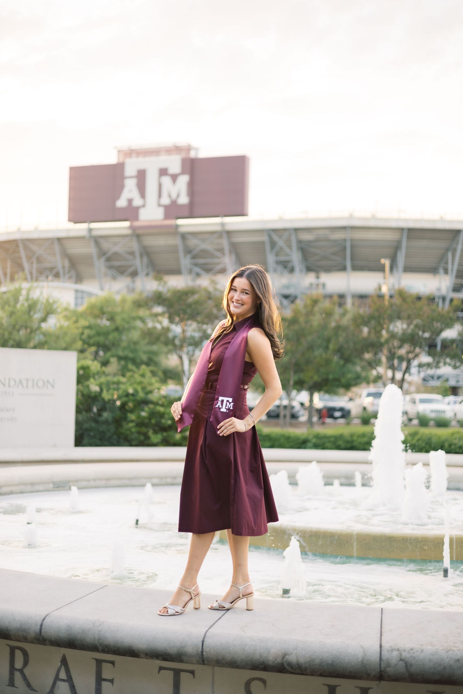 Woman in maroon Texas A&M graduation dress standing by fountain with stadium in background.