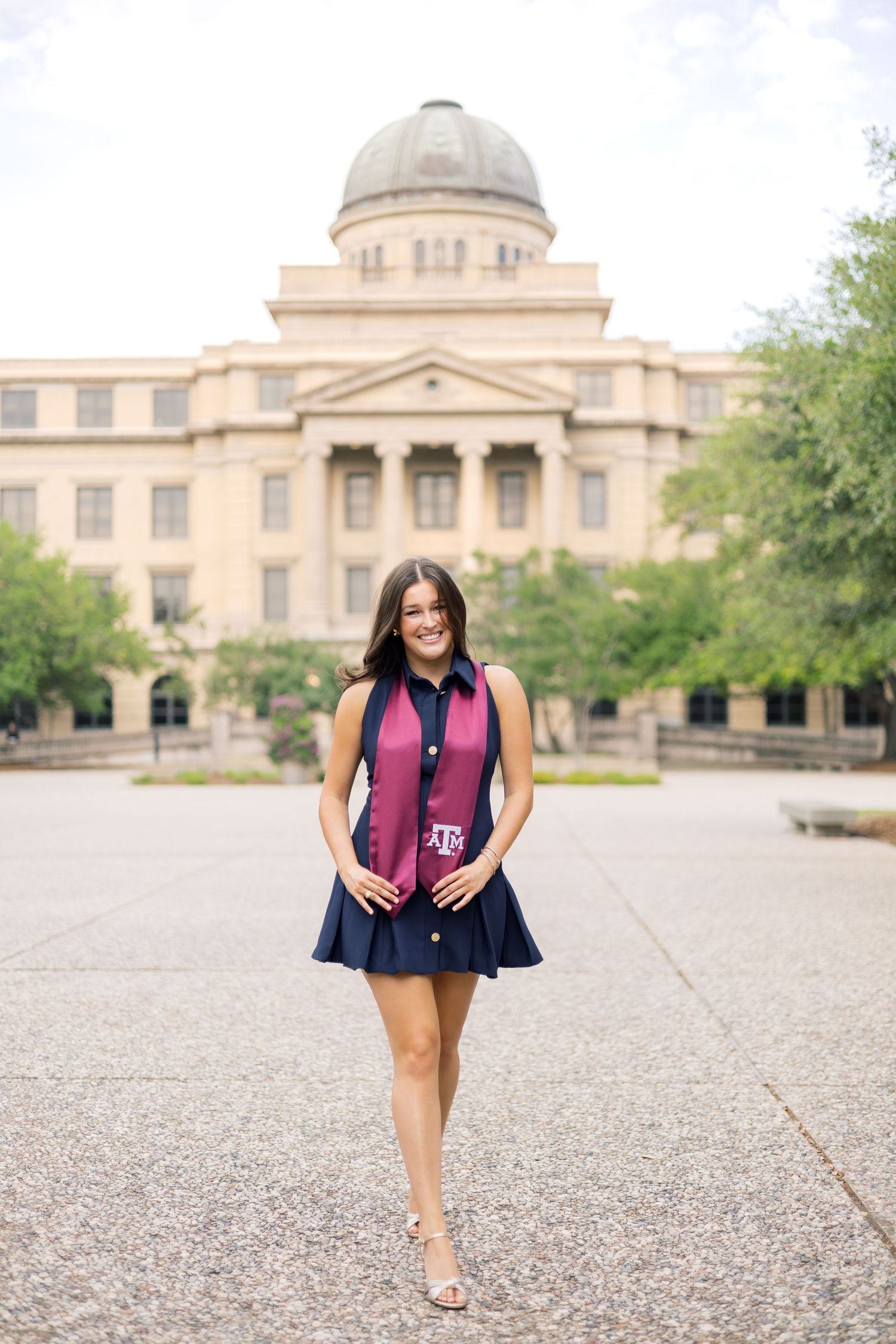 Texas A&M senior in navy dress and maroon stole walks in front of the iconic Academic Building.