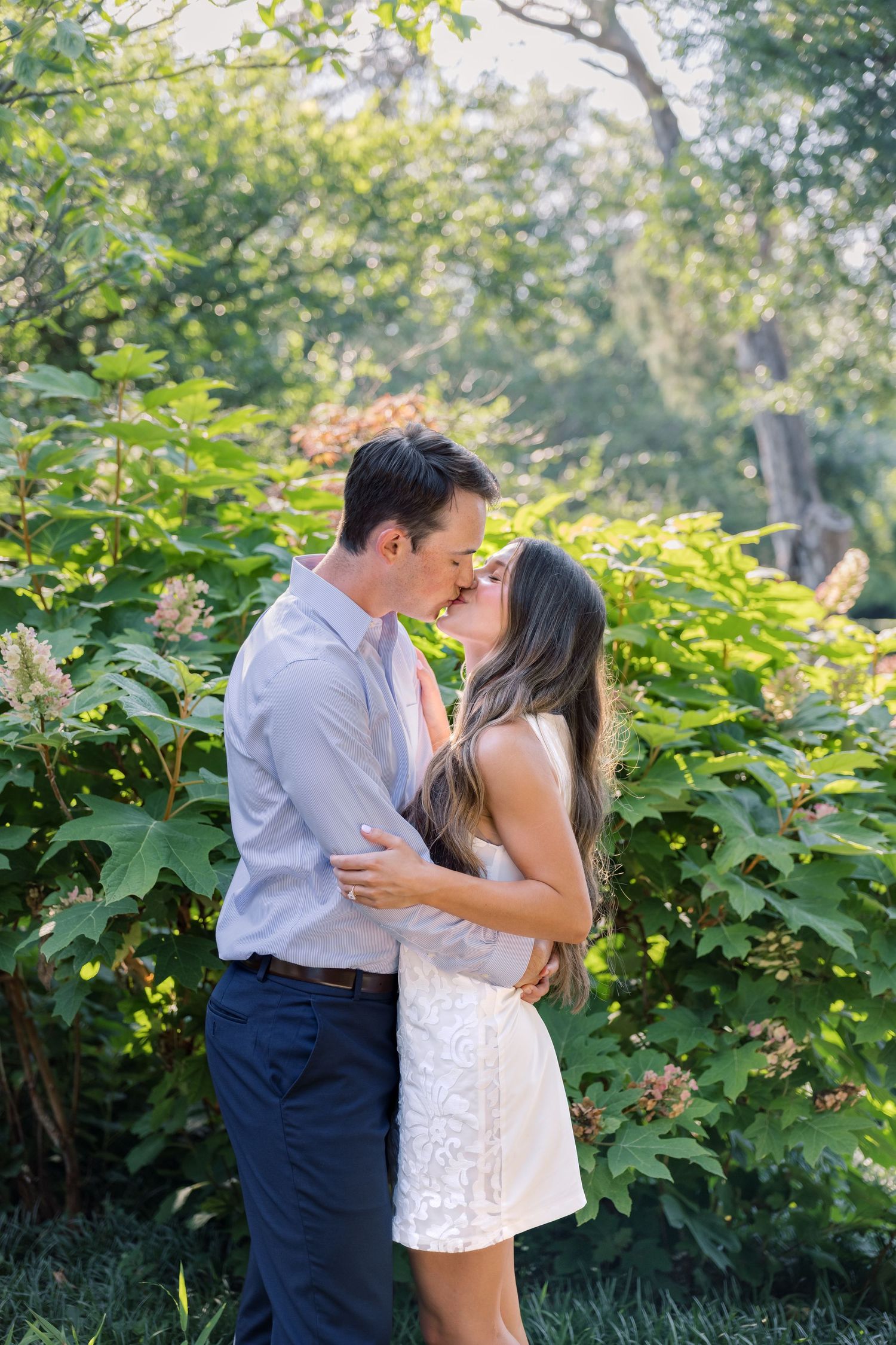 A couple shares a romantic almost-kiss embrace in a lush green garden, man in gray shirt and woman in white lace dress.