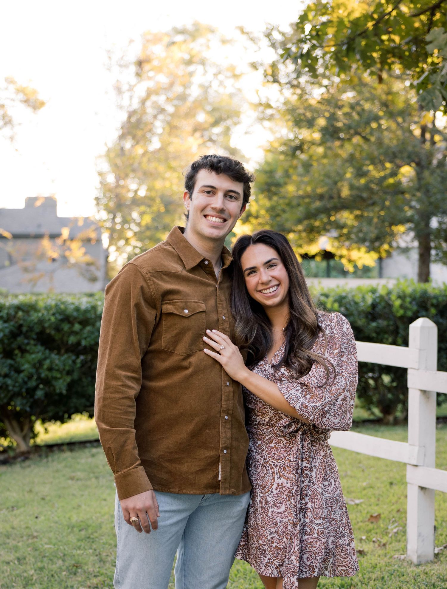 Smiling couple posing outdoors near a white fence with green trees in the background on a sunny day.