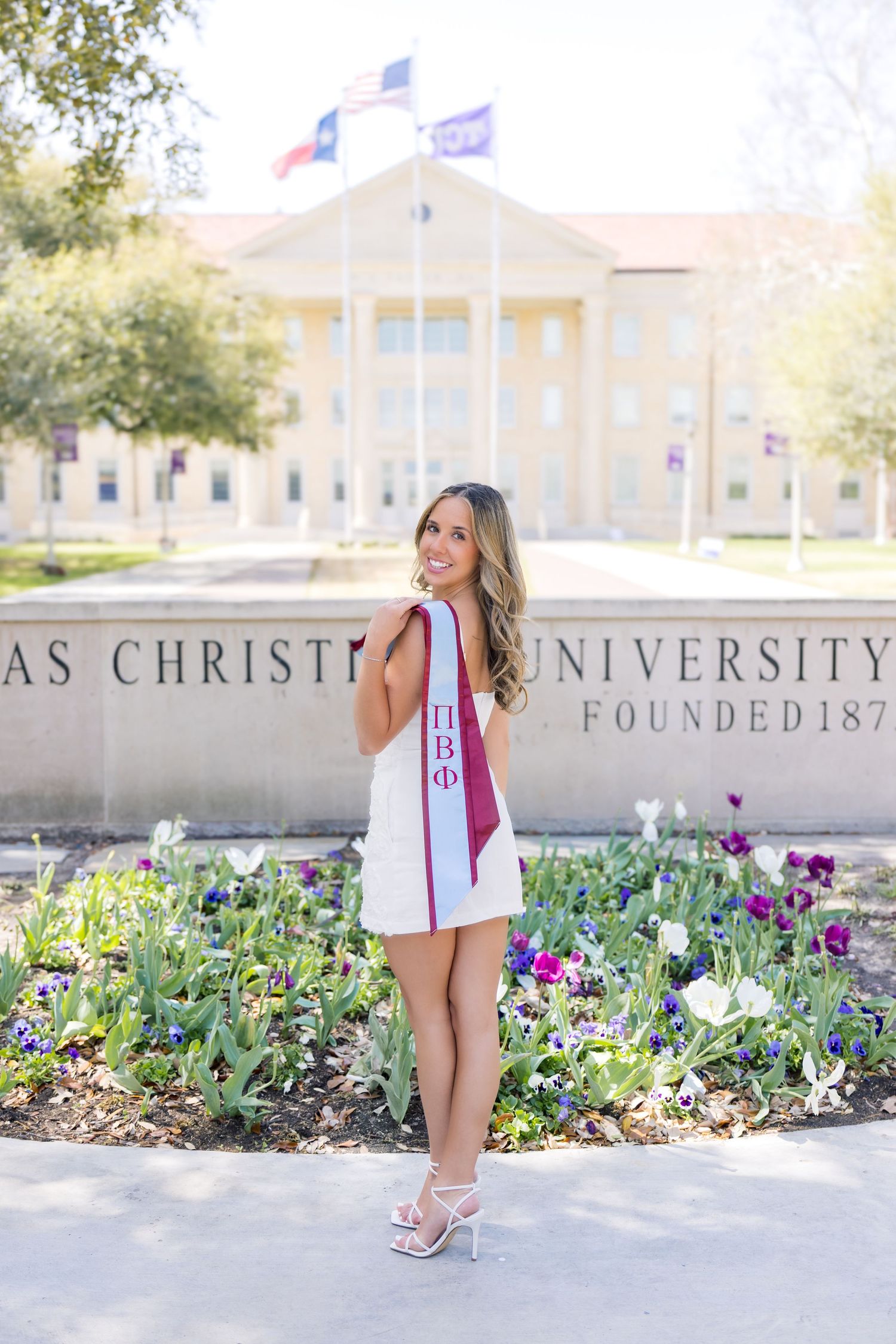Young woman in white dress wearing Pi Beta Phi sash poses at Texas Christian University sign during graduation photos.