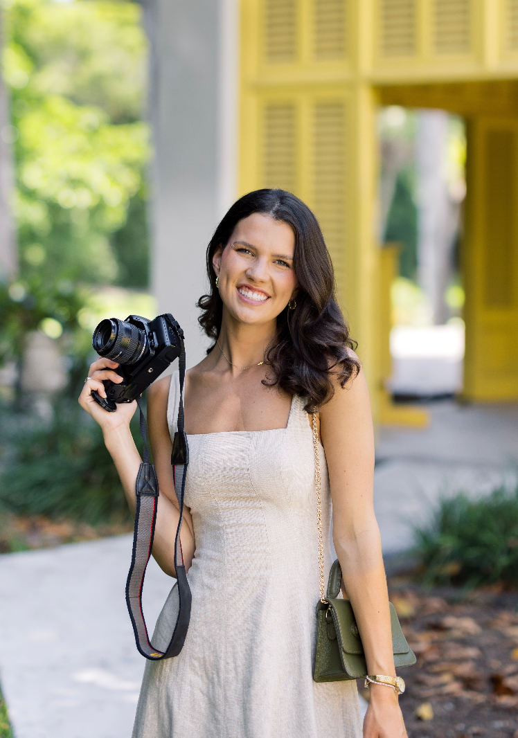 Smiling brunette woman holding a DSLR camera in a white sundress outdoors near a yellow building.