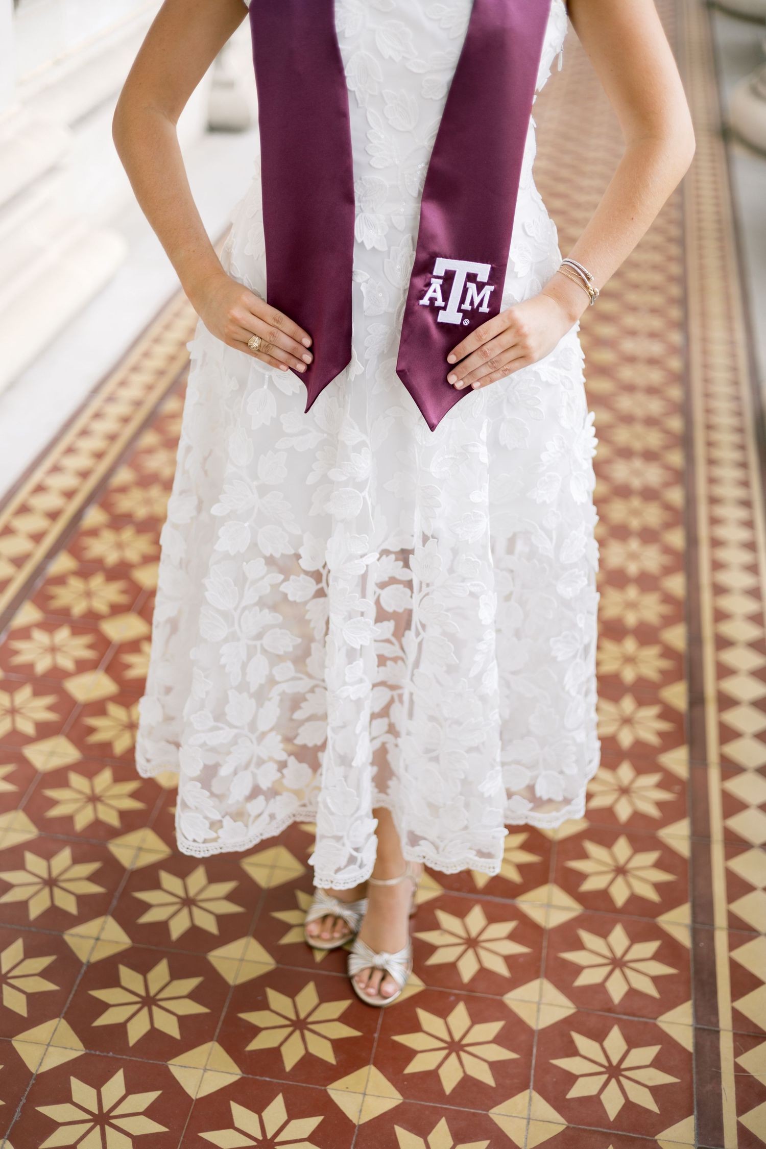 Close-up of Texas A&M graduate in white lace dress holding maroon ATM stole on decorative tiled floor.