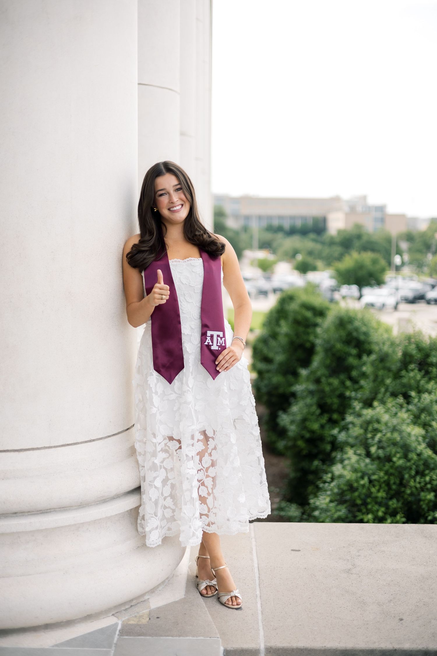 Smiling woman in white lace dress with Texas A&M stole giving thumbs up while leaning against large column outdoors.