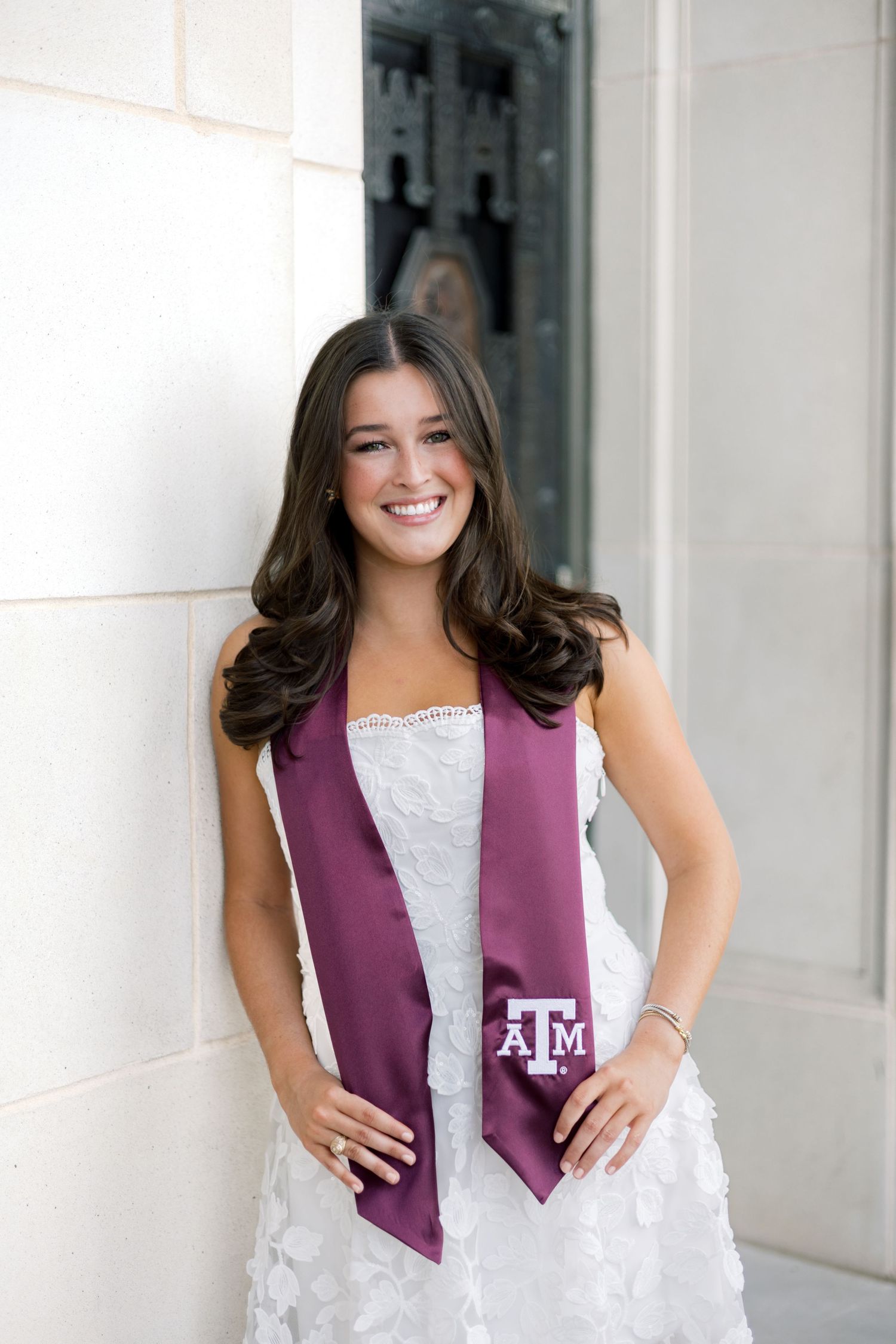 Happy Texas A&M graduate poses in white lace dress with maroon ATM graduation stole against white column.