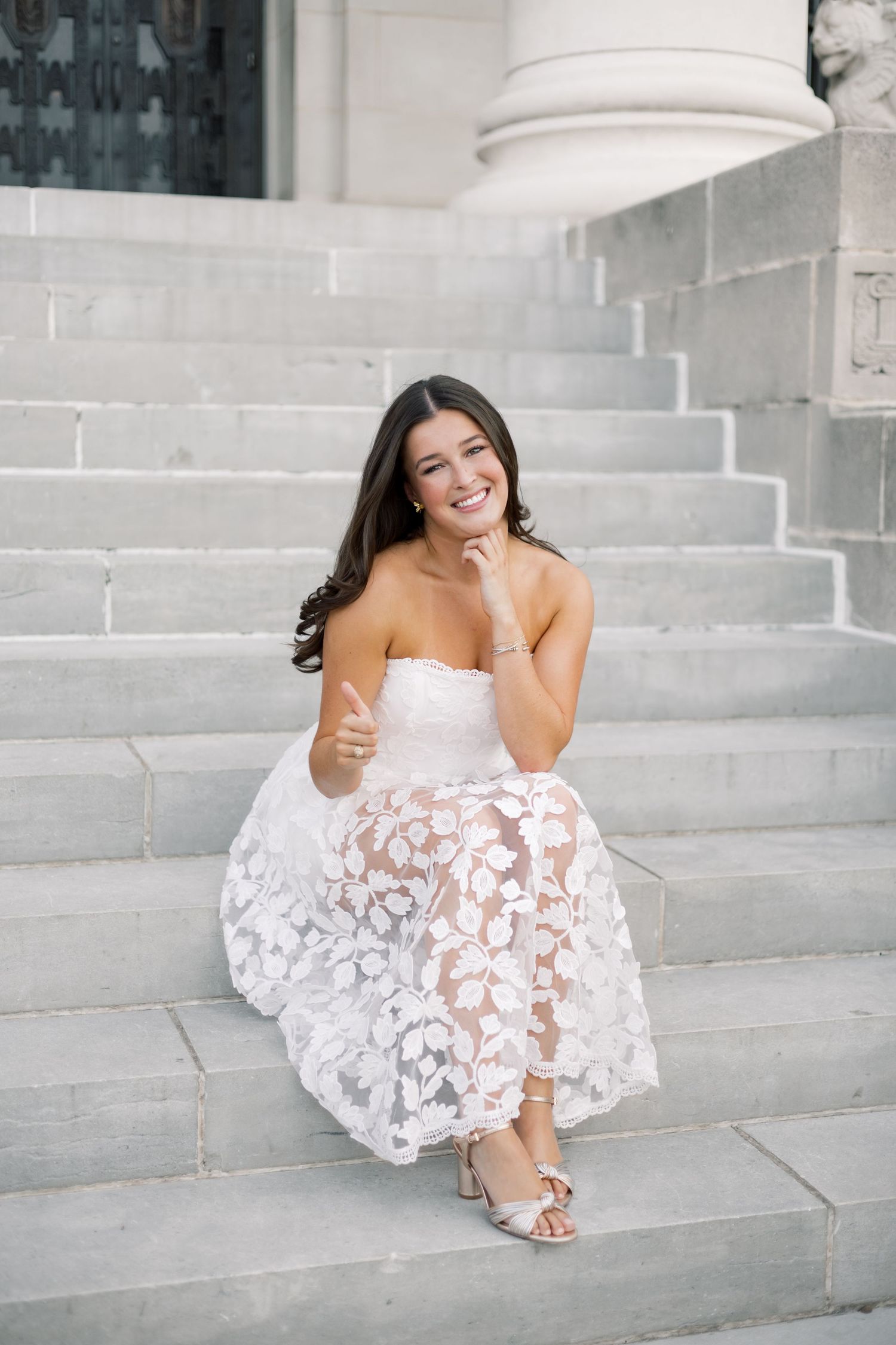 Laughing woman in strapless white lace dress giving thumbs up while sitting on stone steps.