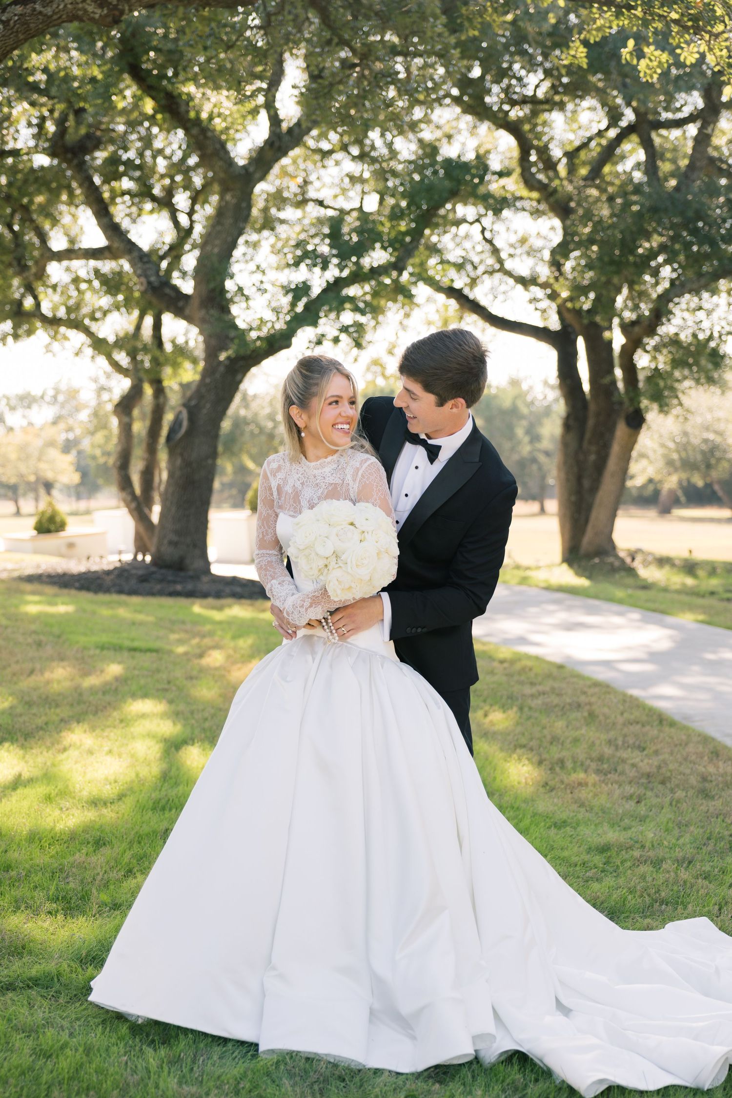 Bride and groom pose under oak trees with white bouquet on wedding day.