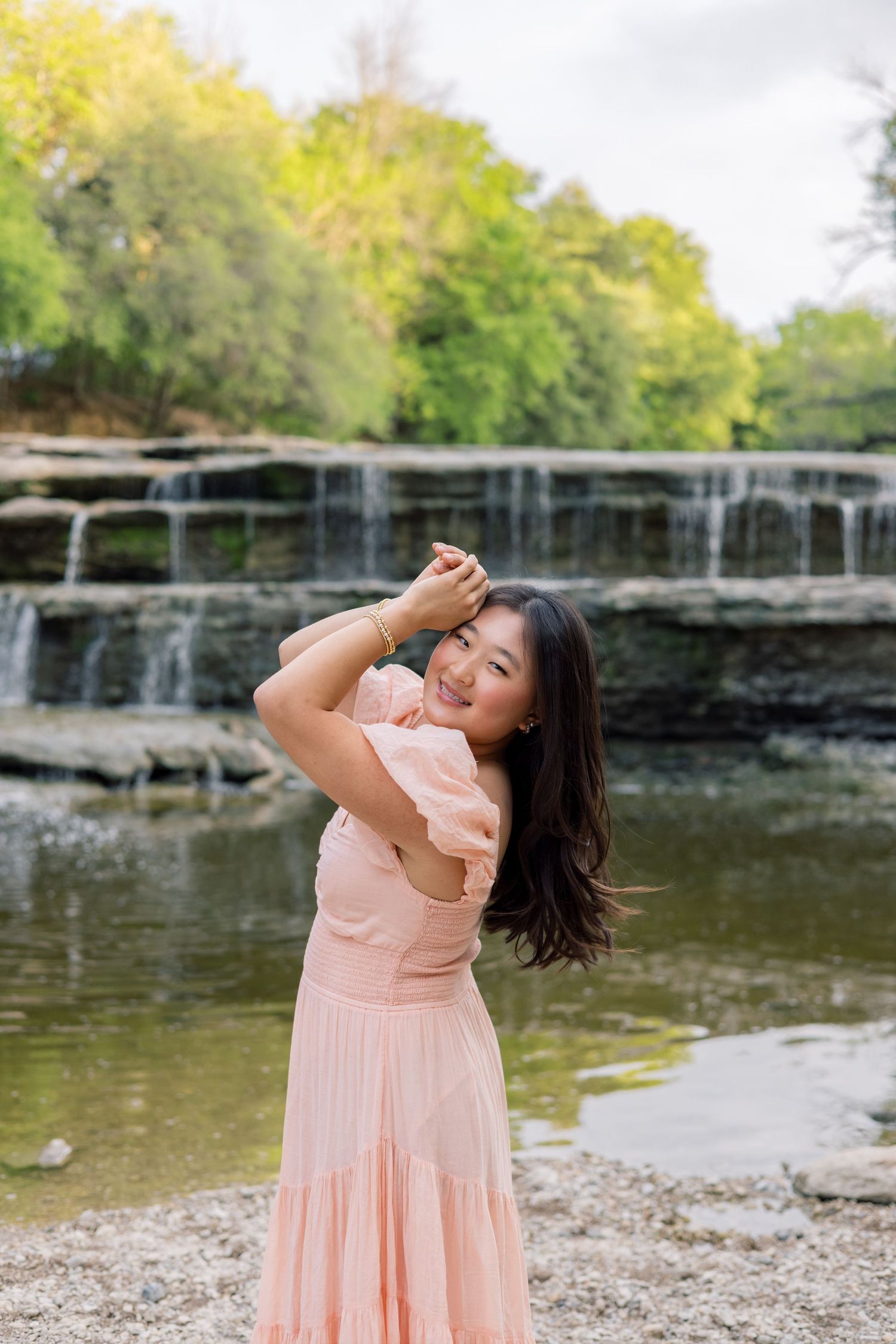 Young woman in pink dress posing near a scenic waterfall and pond in a lush park.