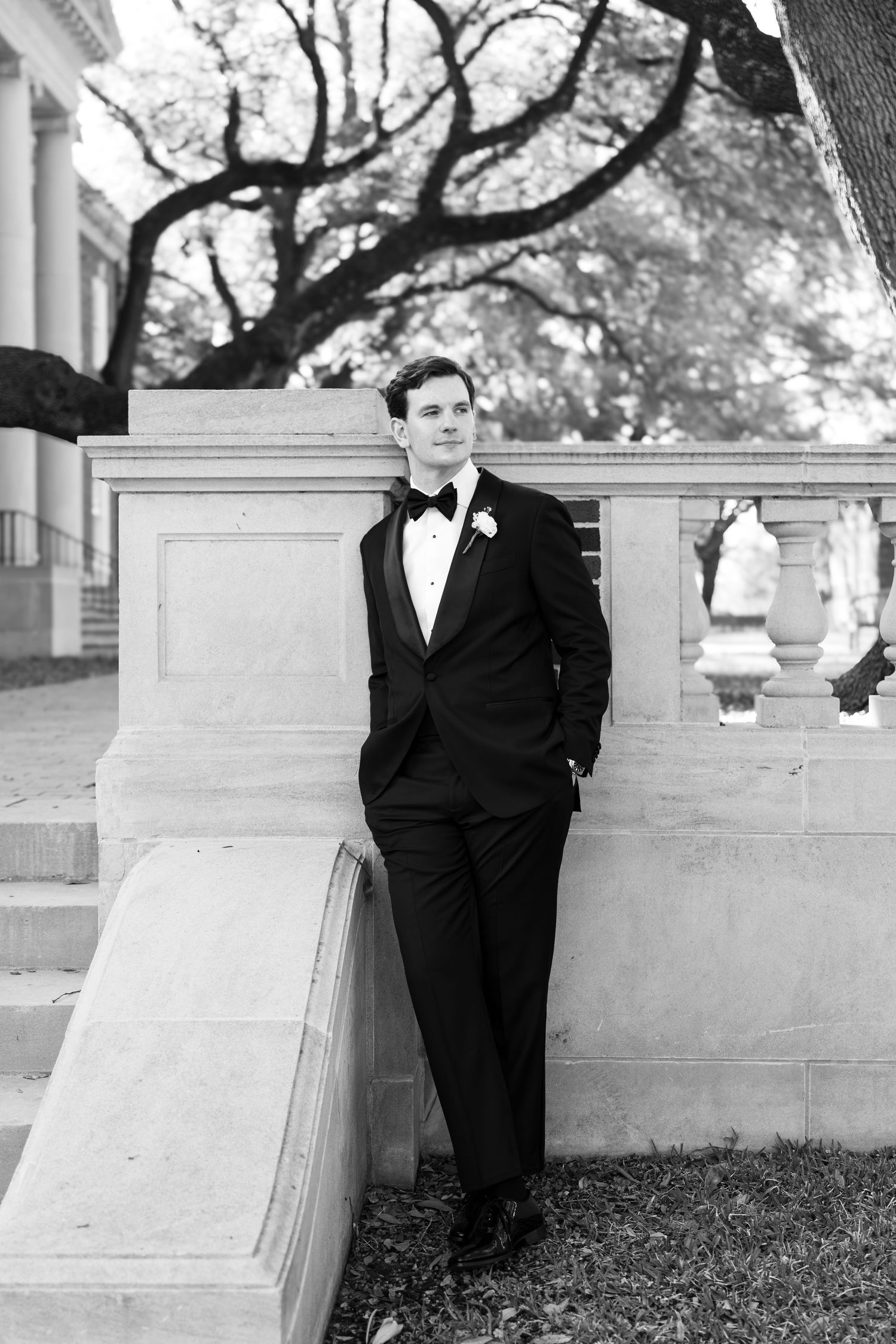 Dapper groom in black tuxedo with boutonniere poses against stone balustrade in black and white.
