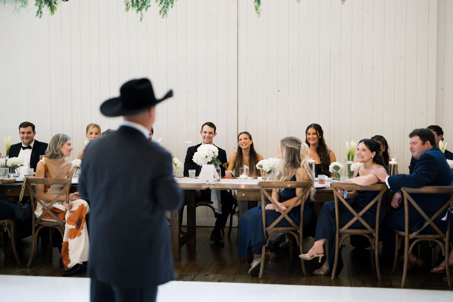 A man in a black cowboy hat addresses elegantly dressed wedding guests seated at a decorated table in a bright venue.