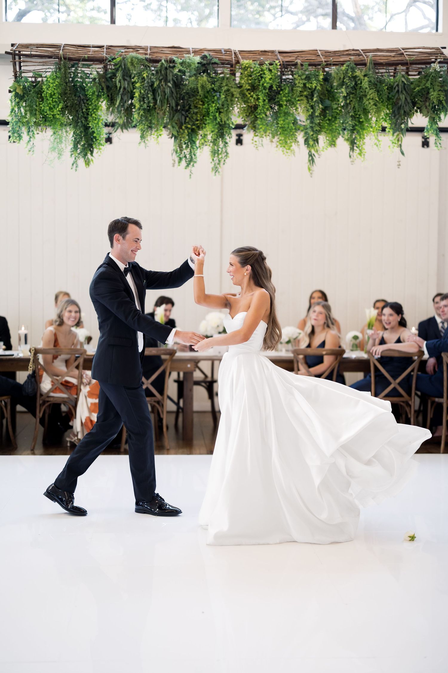 Bride and groom share their first dance under lush greenery installation at elegant wedding reception.