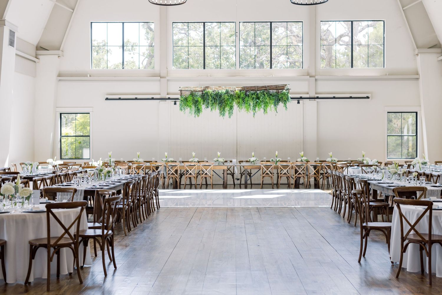 Elegant wedding reception hall with white walls, wooden floors, long tables, cross-back chairs, and lush green floral ceiling installation.