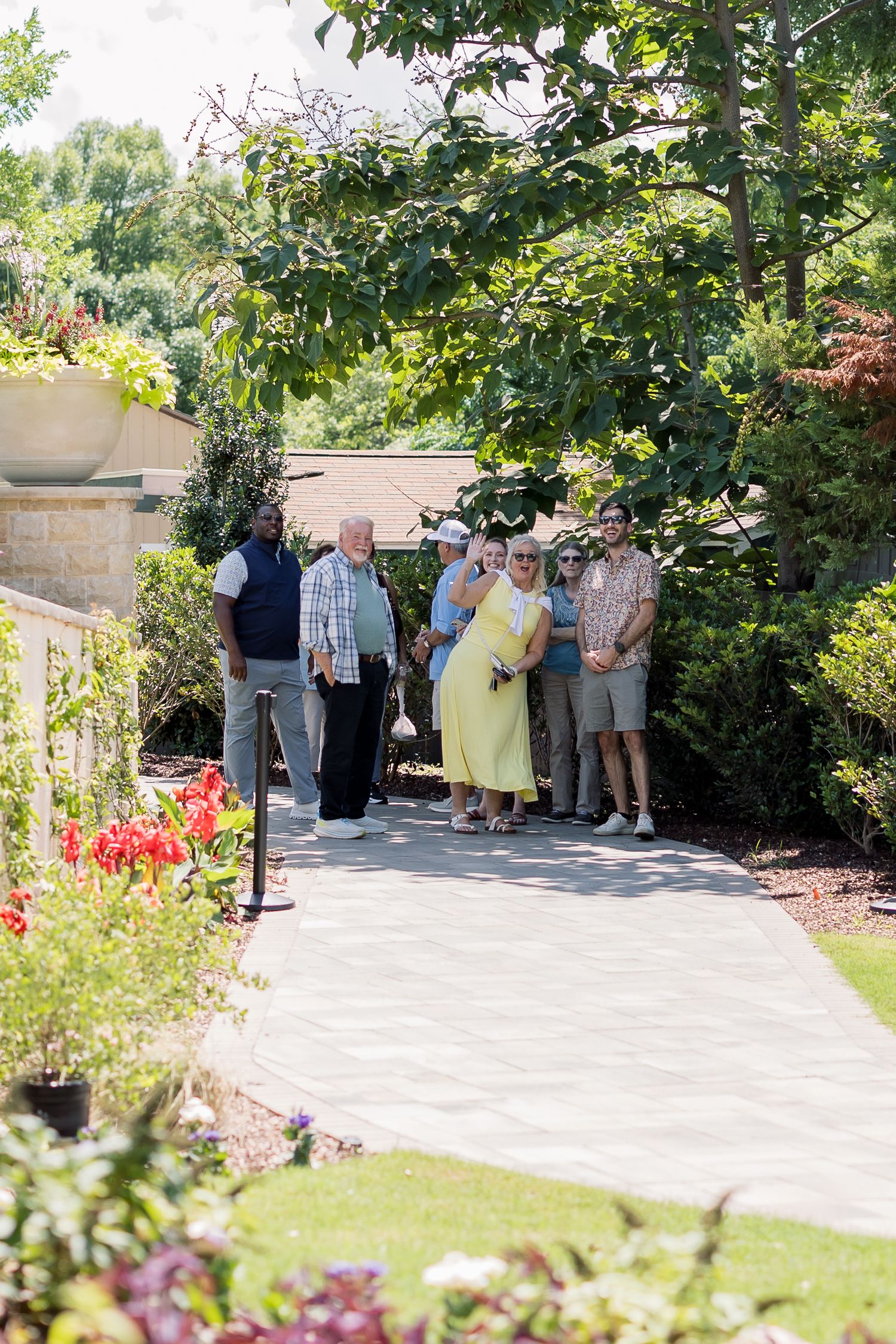 Group of people posing on a garden pathway surrounded by lush green trees and colorful flowers on a sunny day.