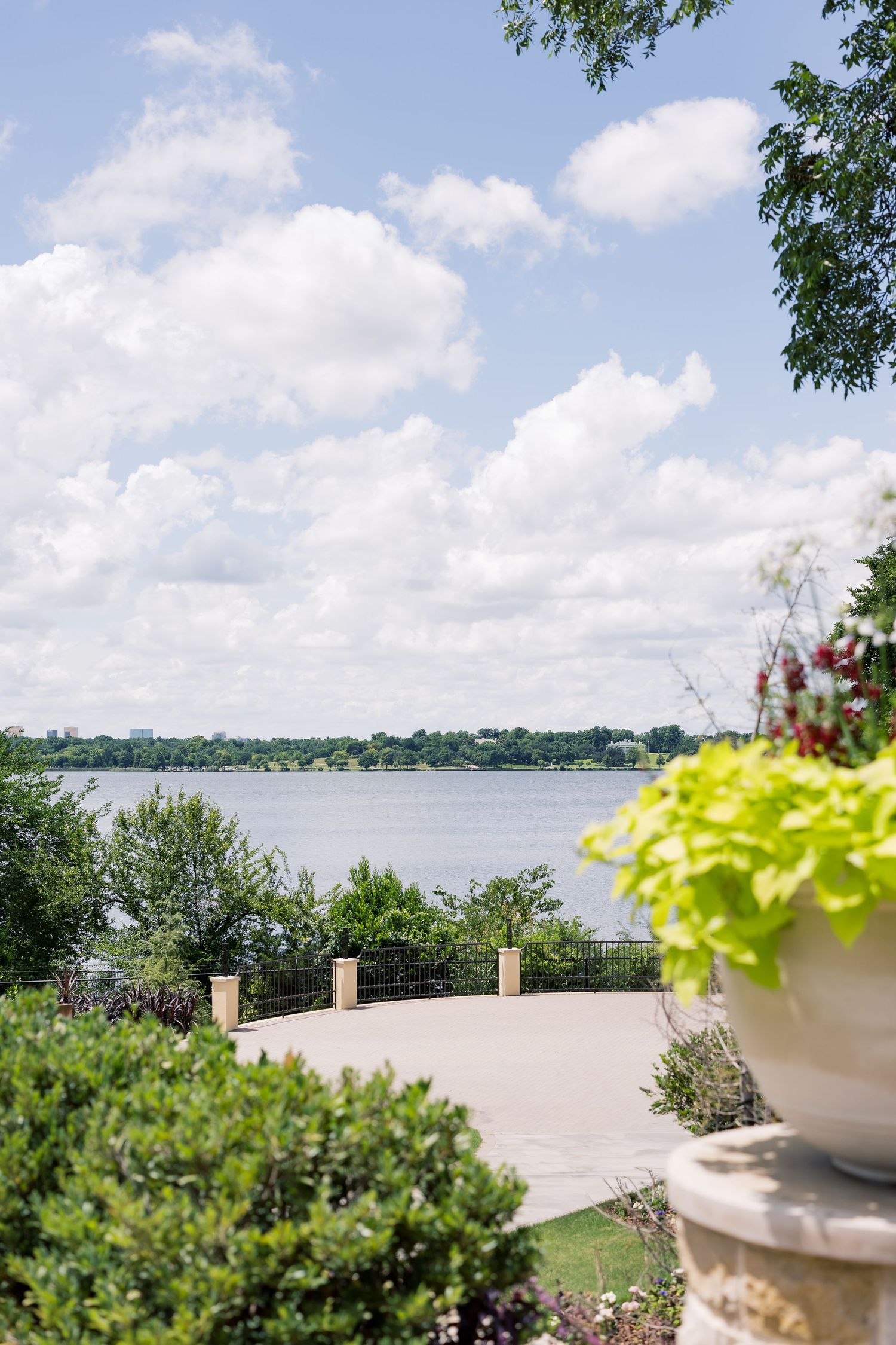Scenic lakeside patio with lush greenery, decorative stone planter, and cloudy blue sky reflecting over calm water.