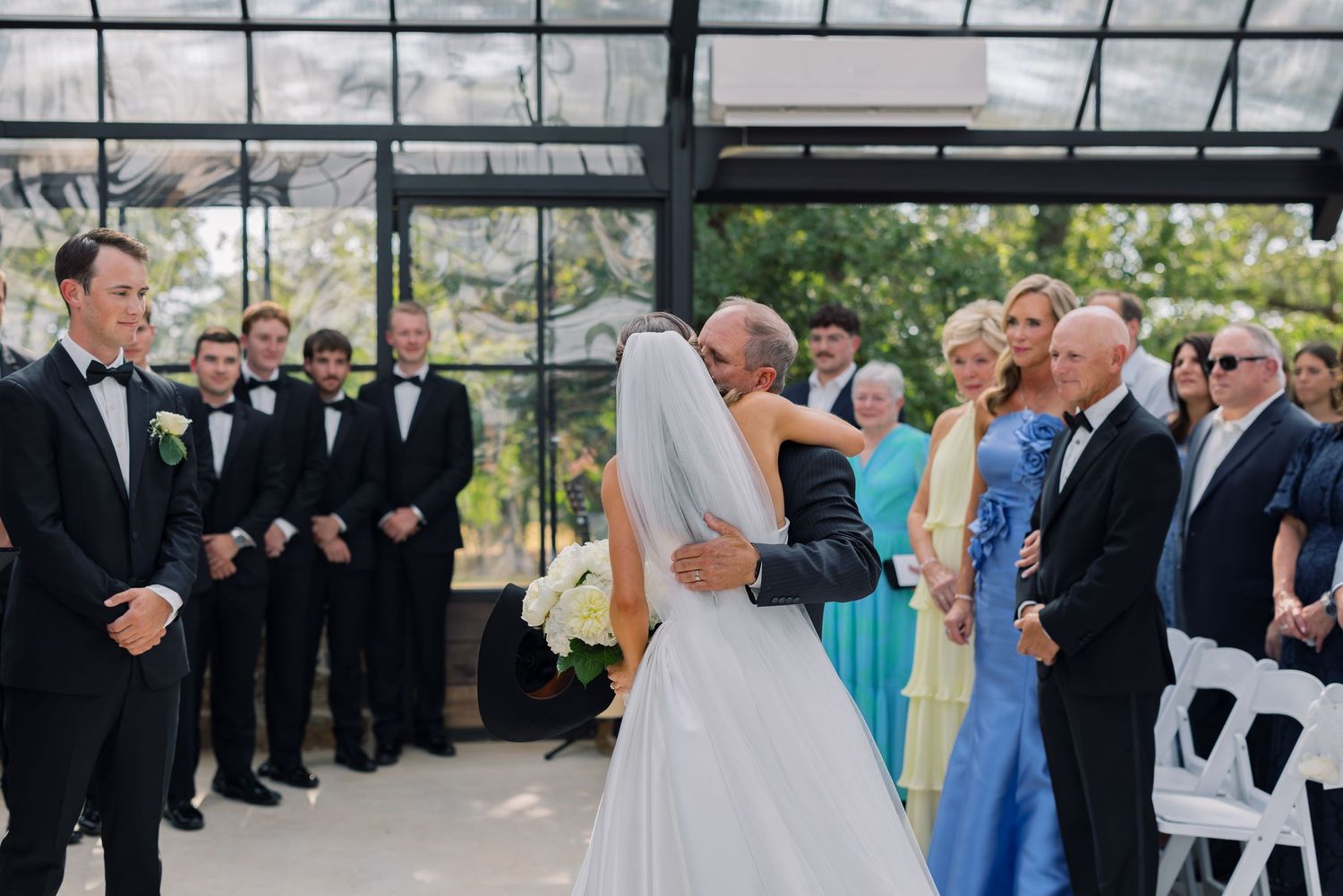 Bride and groom share first dance at elegant greenhouse wedding ceremony with bridal party and guests watching.
