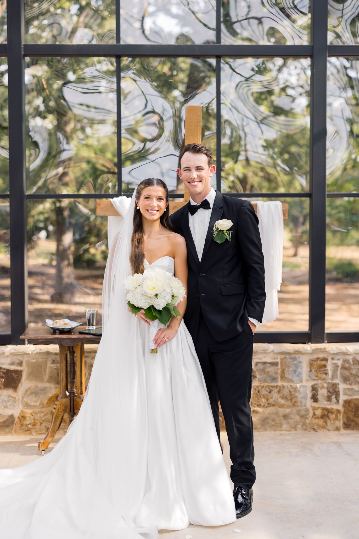 Bride in white strapless gown holding white bouquet stands beside groom in black tuxedo before large glass windows.