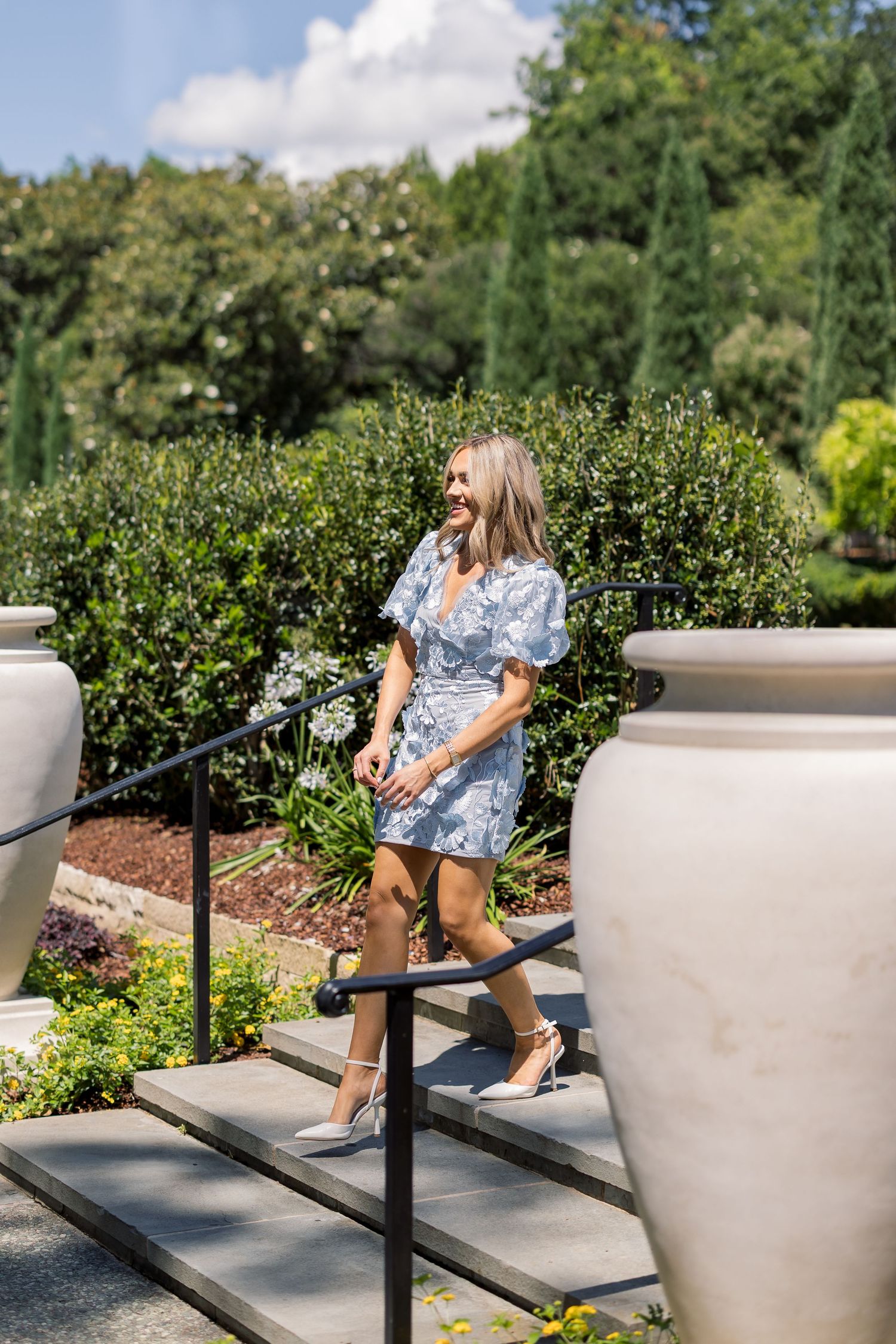 Woman in blue floral mini dress and heels walking down garden steps surrounded by large stone urns.