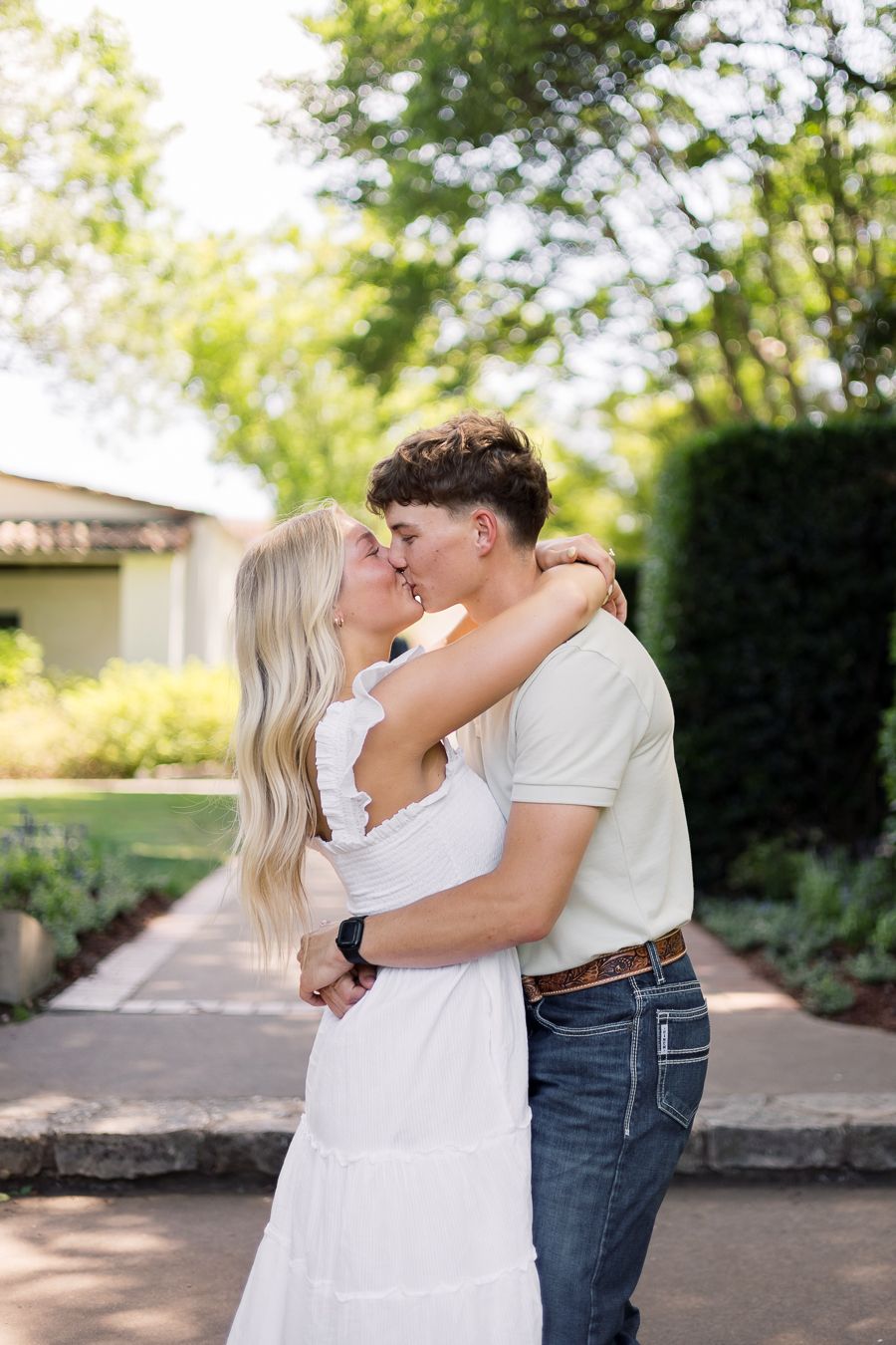 A couple shares a romantic kiss outdoors in a lush garden, woman in white dress, man in jeans and cream shirt.