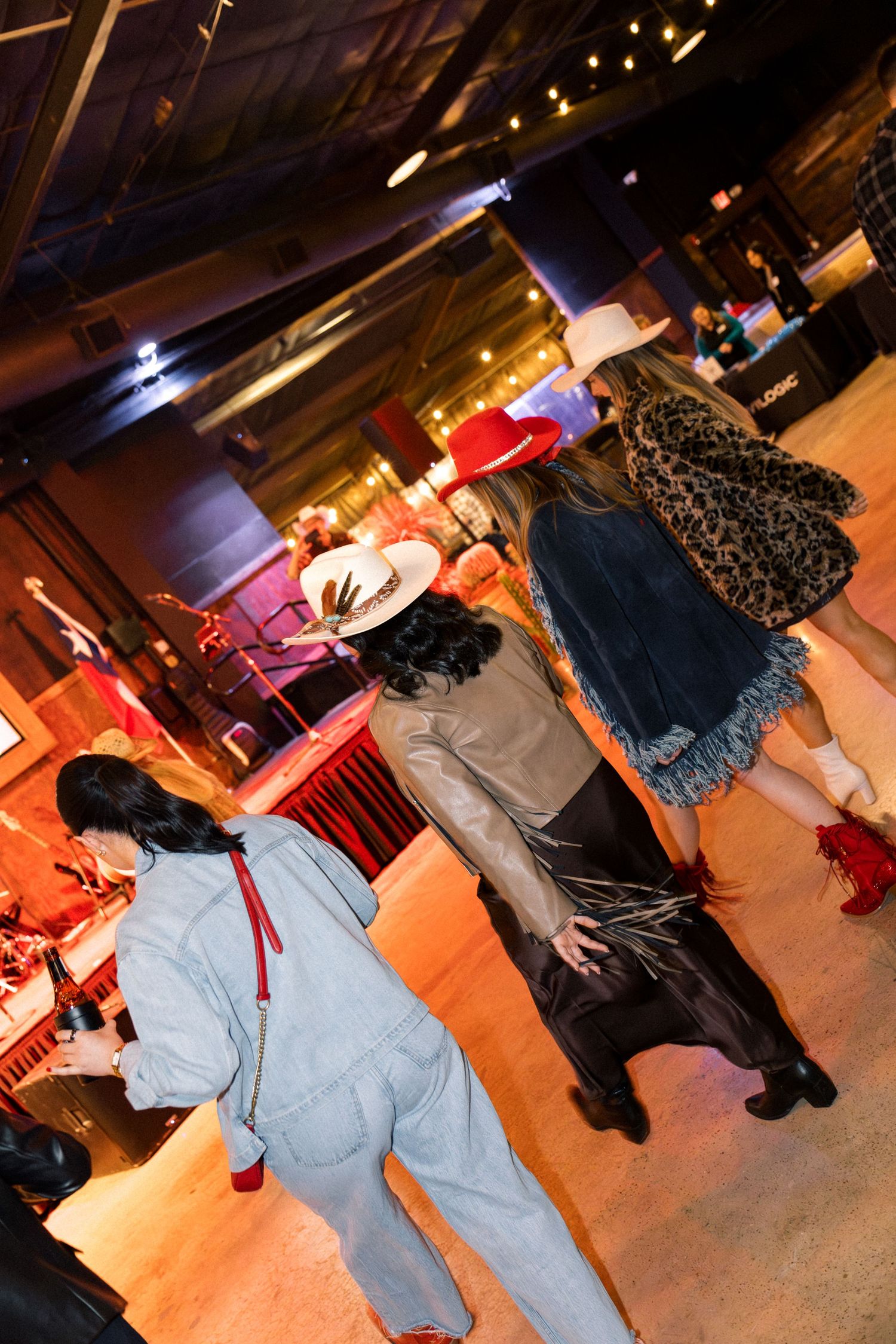 Three women in cowboy hats and western outfits walk through a vibrant indoor venue with warm lighting.