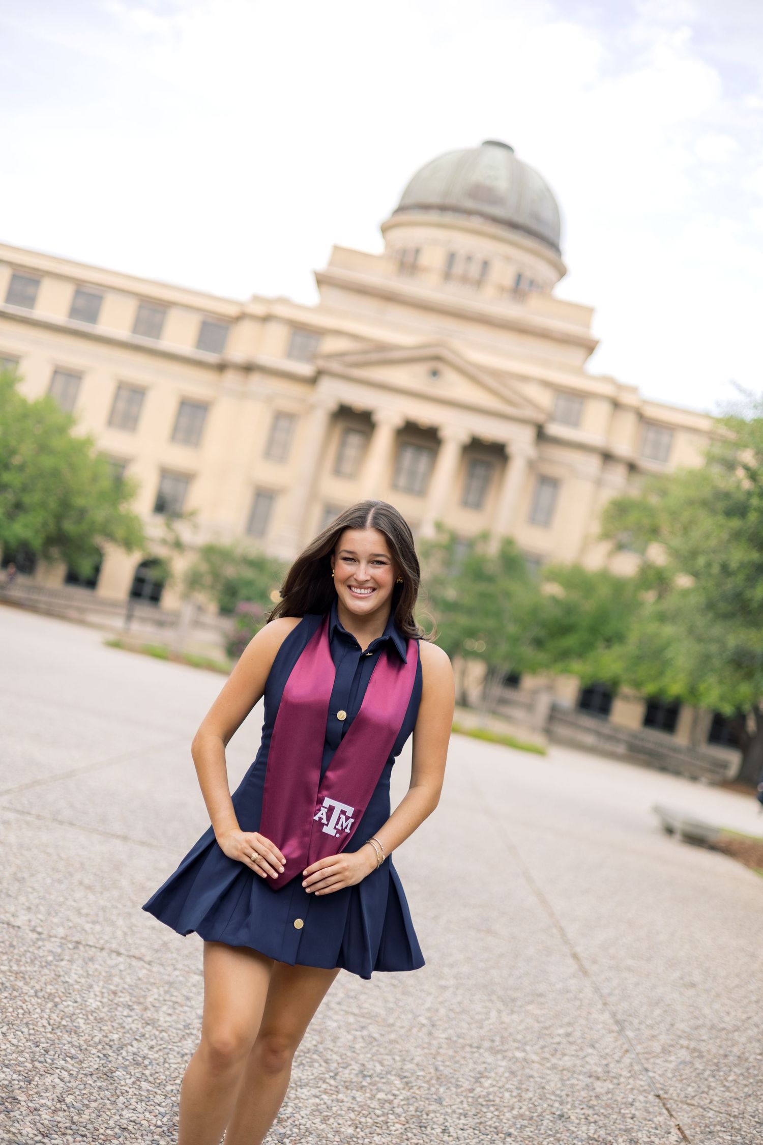 Smiling Texas A&M graduate poses in navy dress and maroon stole with the Academic Building behind her.