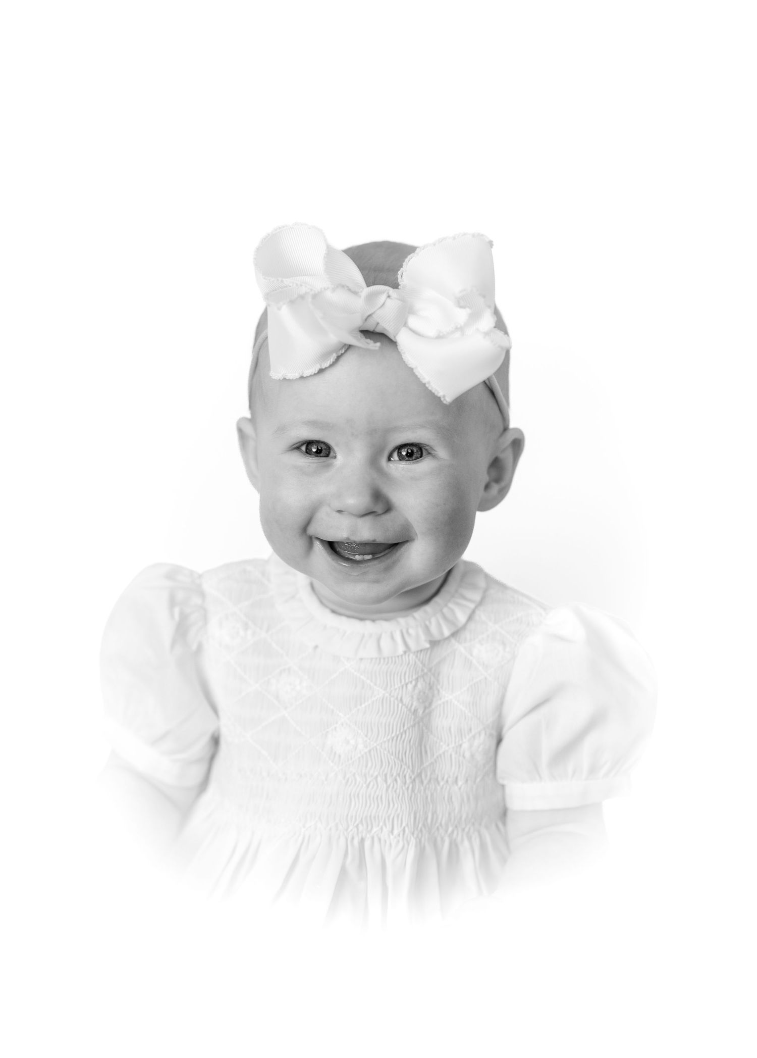 Black and white portrait of a smiling baby girl wearing a white dress and large bow headband.