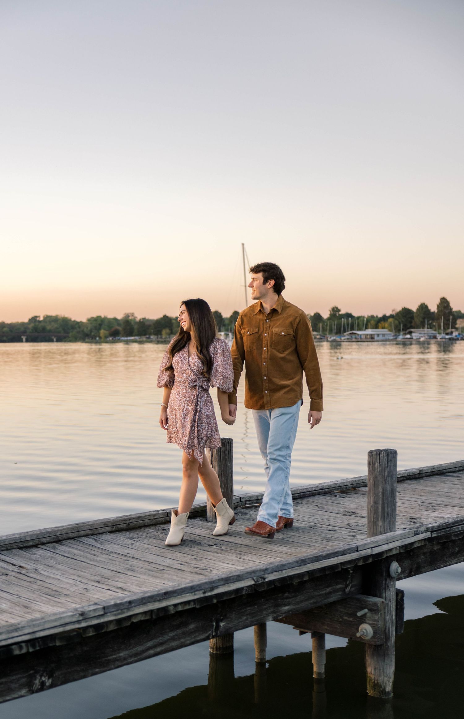 Couple walking hand in hand along a wooden dock at sunset with calm lake and sailboats in background.