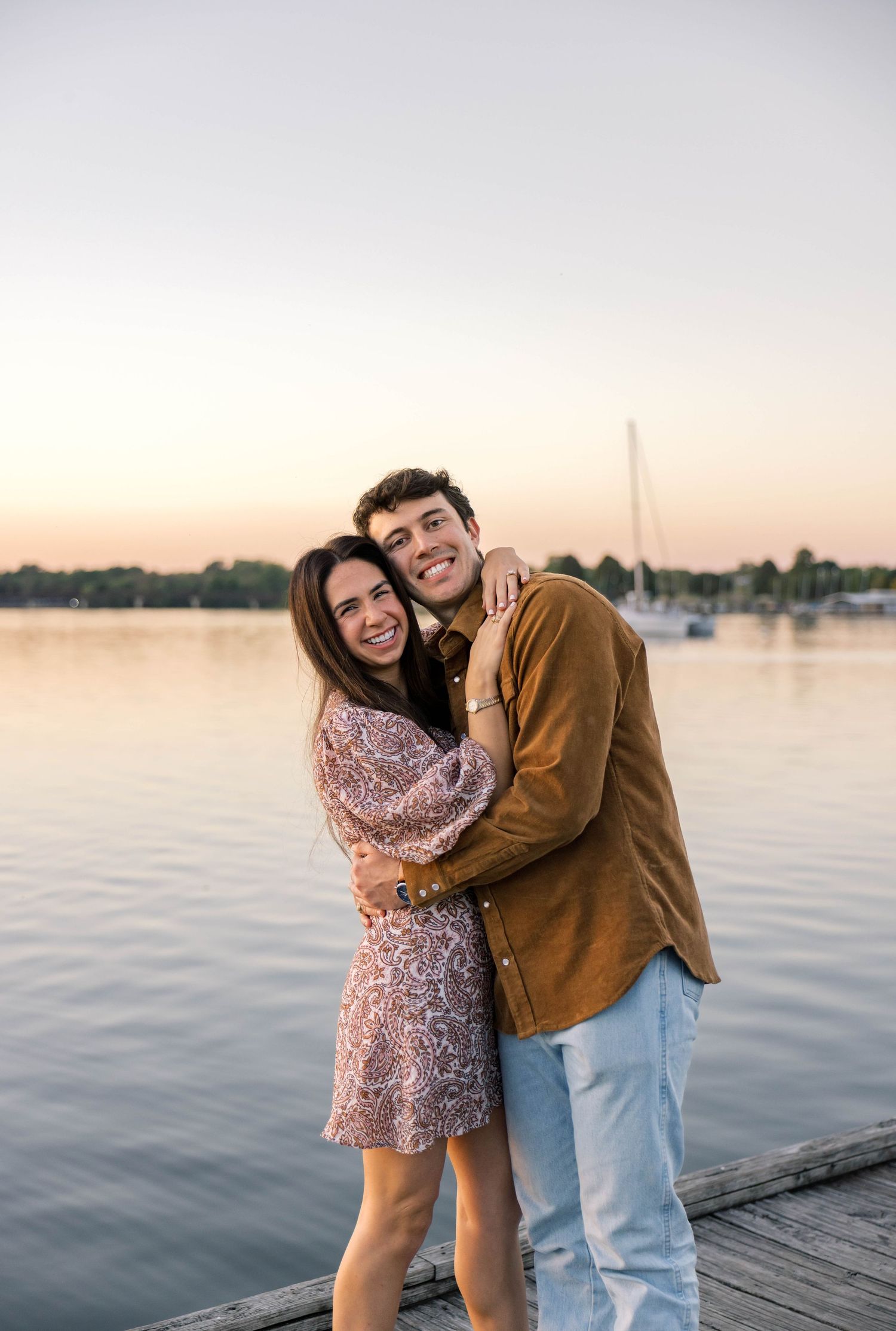 Joyful couple embracing and smiling on a dock at sunset with a calm lake and sailboat behind them.