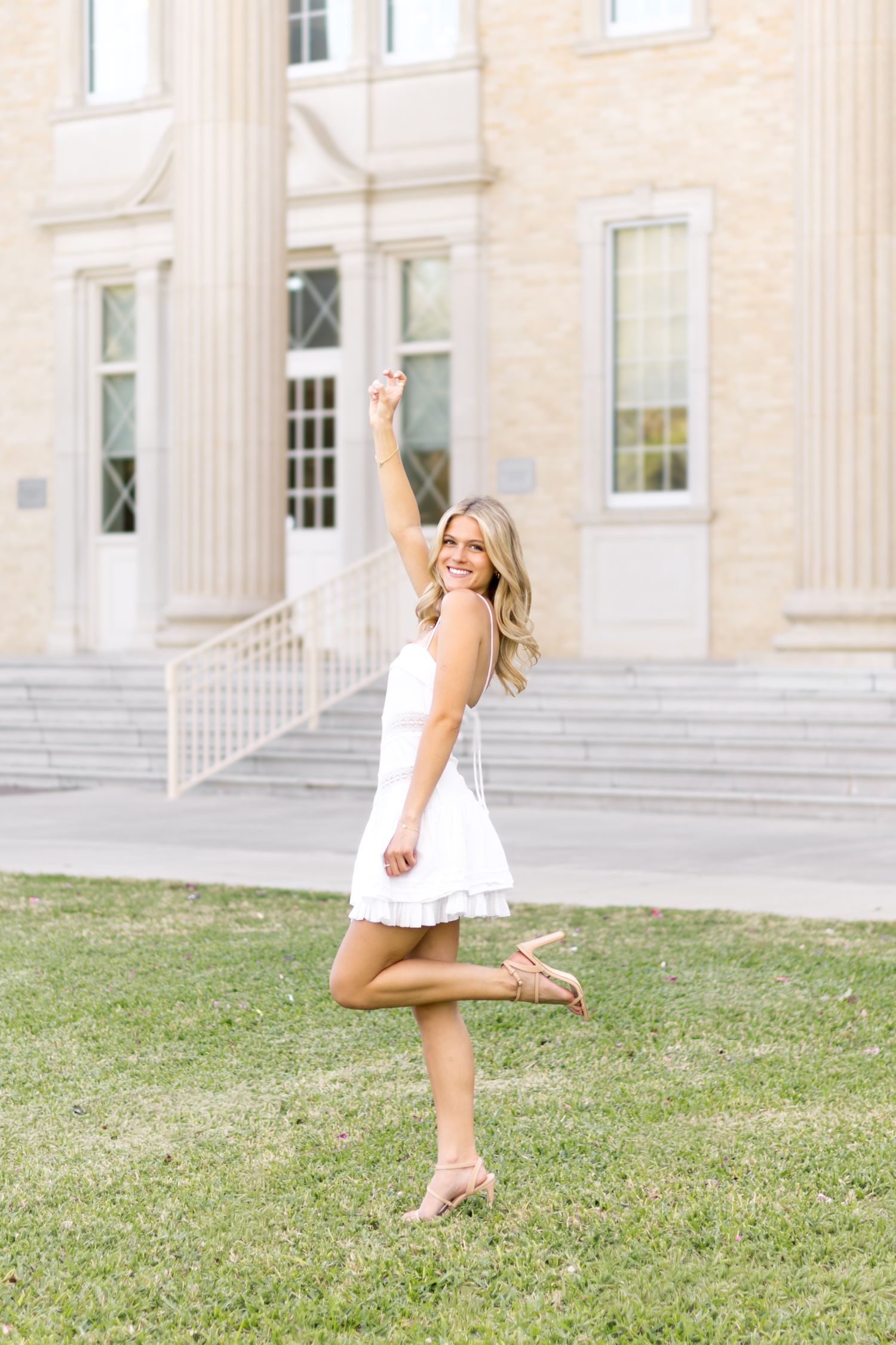 Smiling blonde woman in white mini dress celebrating outdoors near a classic university building.