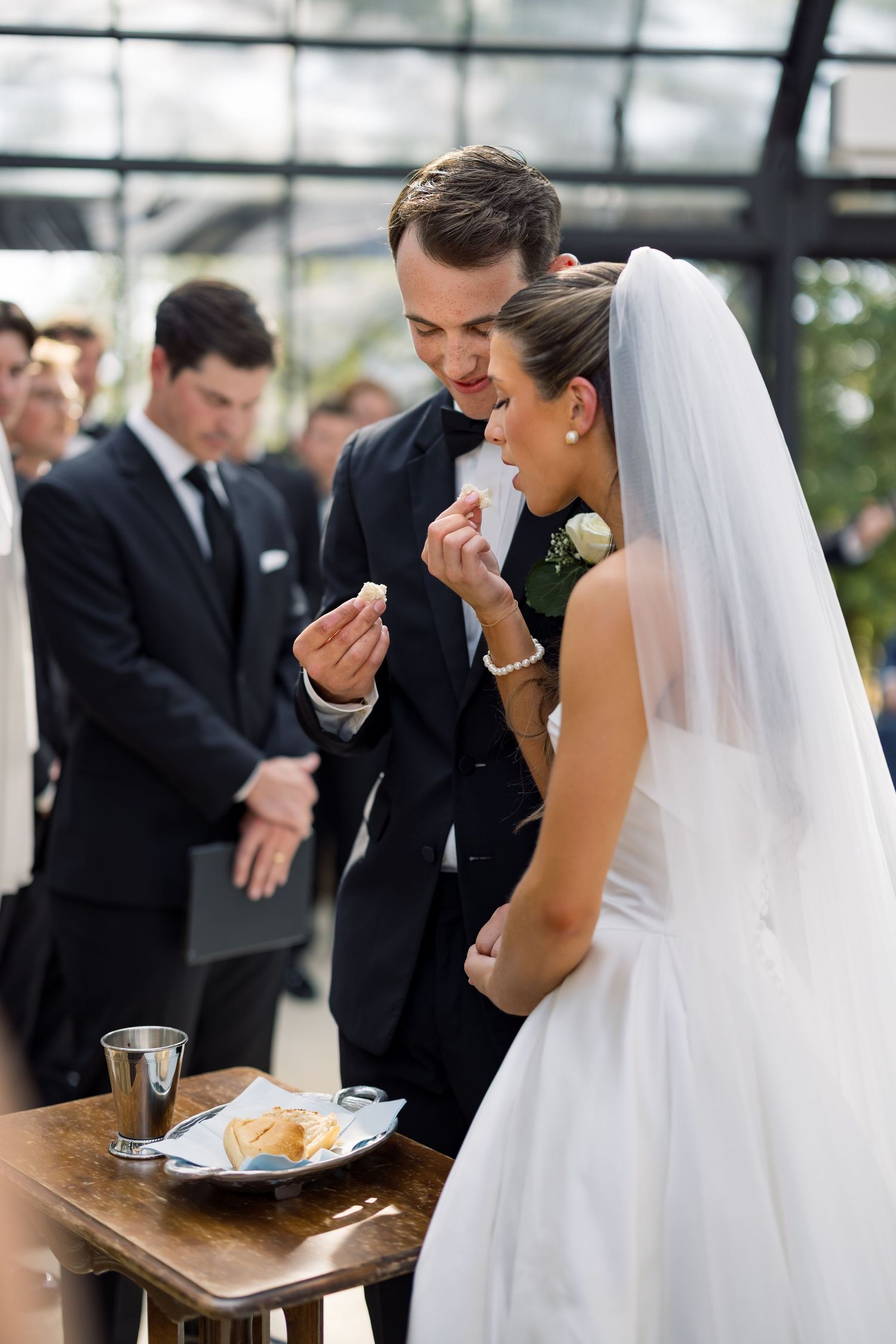 Bride and groom sharing wedding cake at reception ceremony, guests watching in background.
