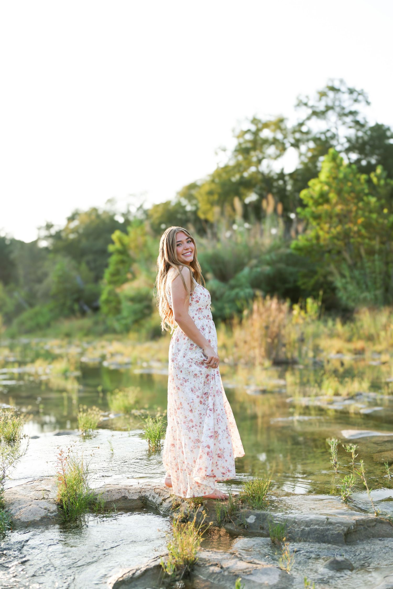 Smiling woman in floral maxi dress standing on rocks near a serene pond surrounded by lush greenery in golden hour light.