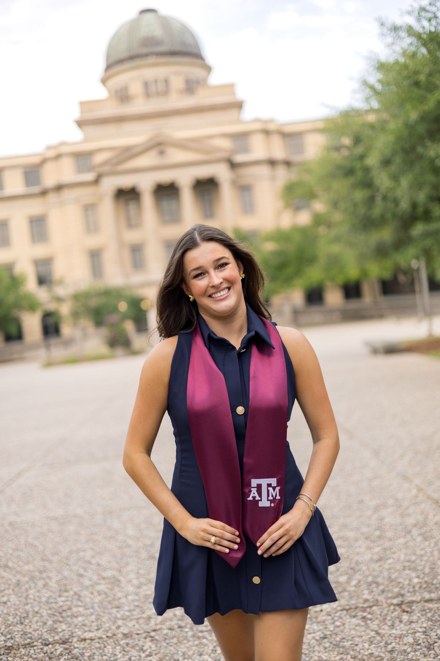 Smiling young woman in navy dress and maroon Texas A&M graduation stole poses in front of campus building.