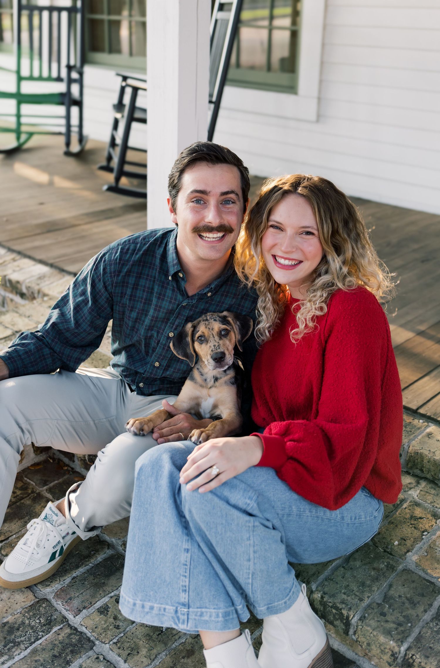 A smiling couple sits on a porch steps holding an adorable puppy, woman in red top and man in plaid shirt.
