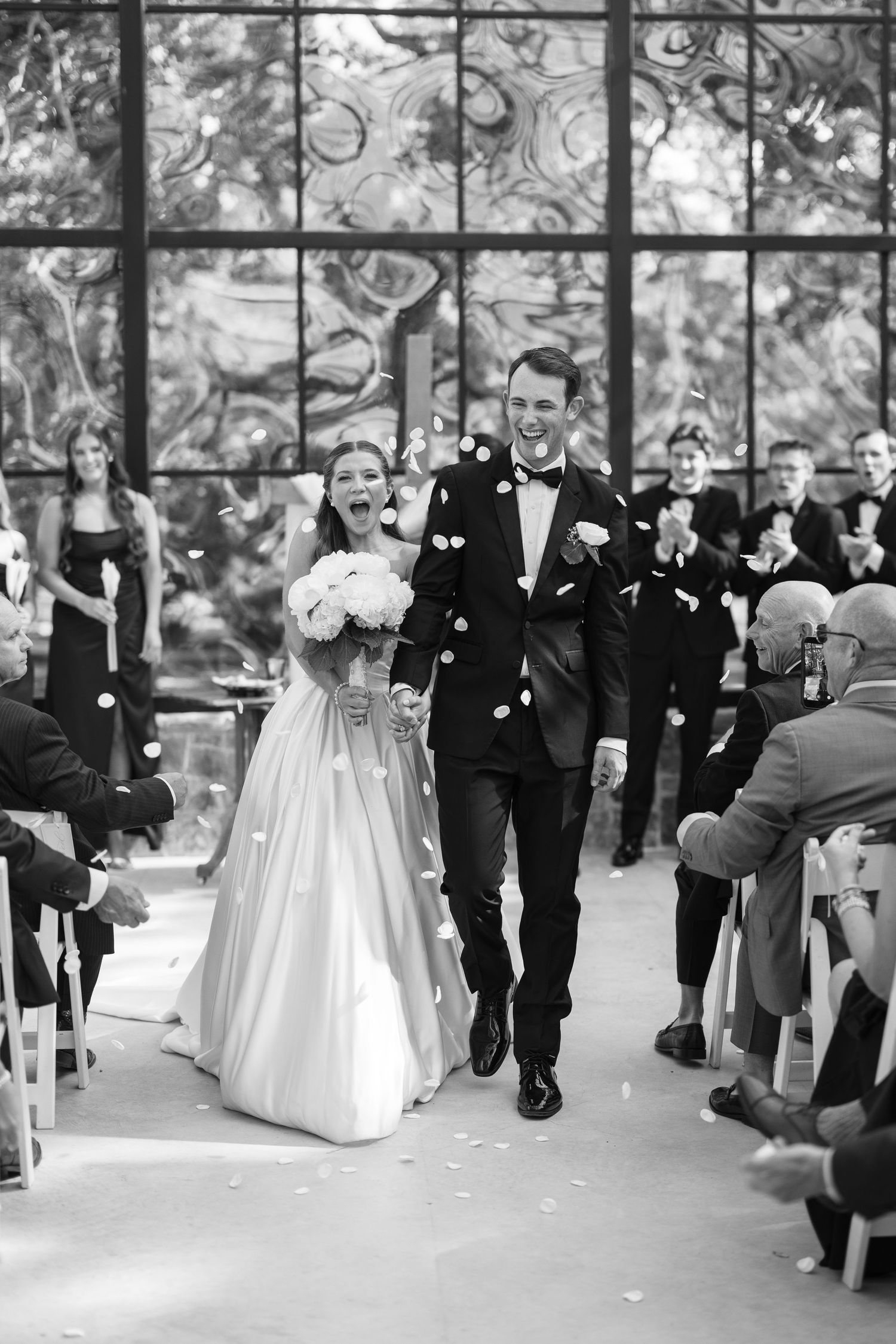 Black and white photo of joyful newlyweds walking down the aisle as guests toss flower petals at their wedding ceremony.