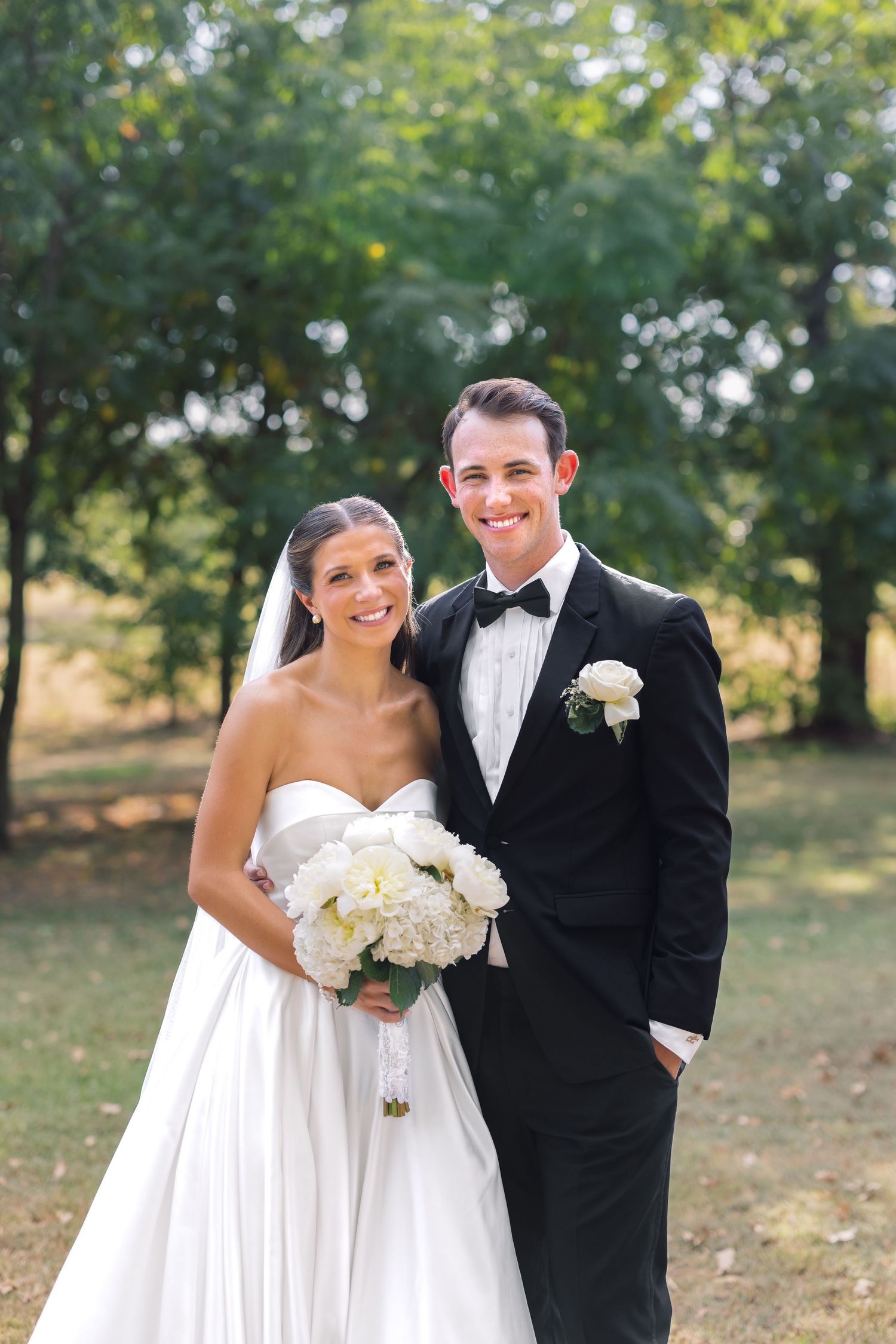 Smiling bride in strapless white gown holding bouquet stands beside groom in black tuxedo outdoors.