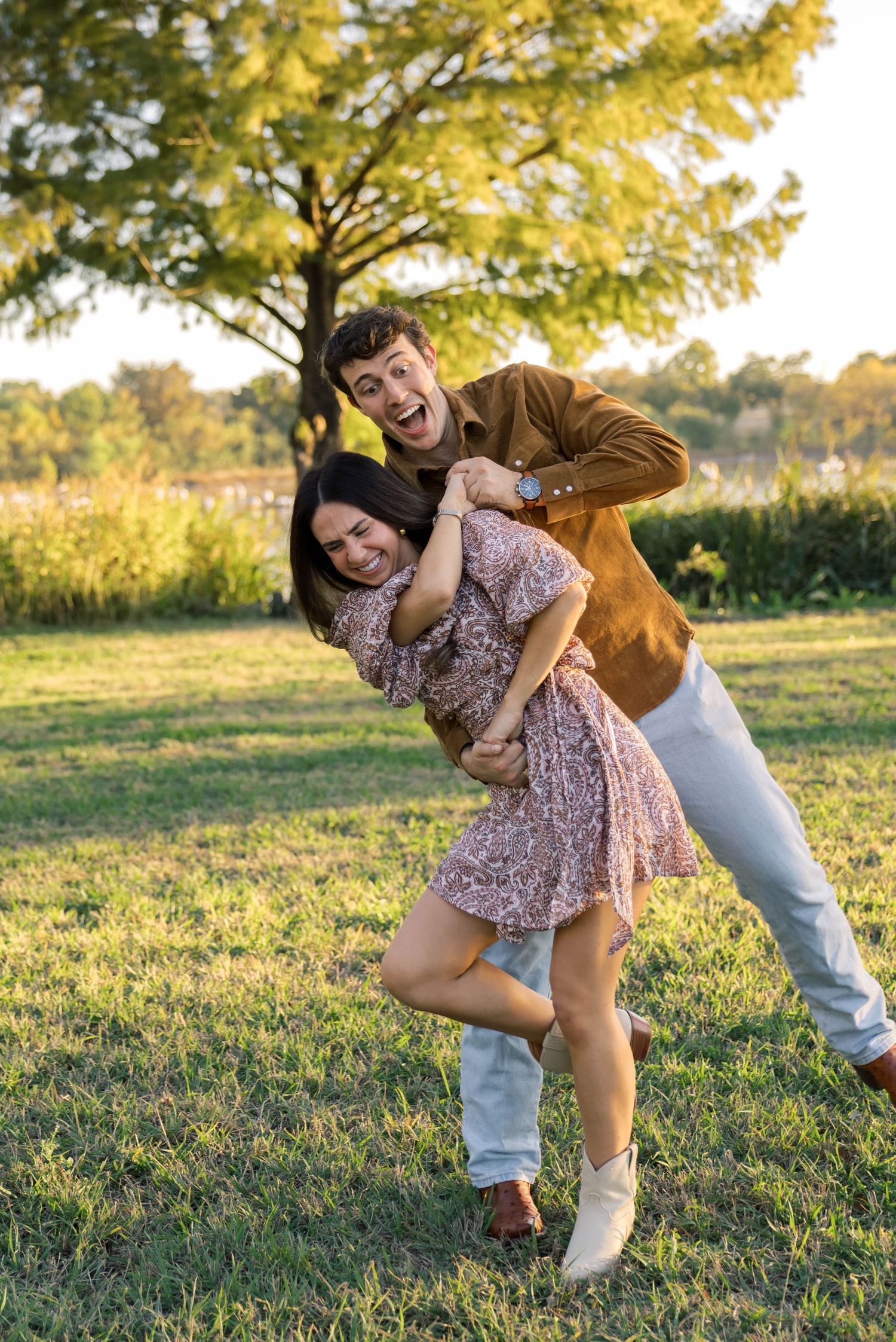 Joyful couple playing in a sunny park, man hugging woman from behind as she laughs in a floral dress.