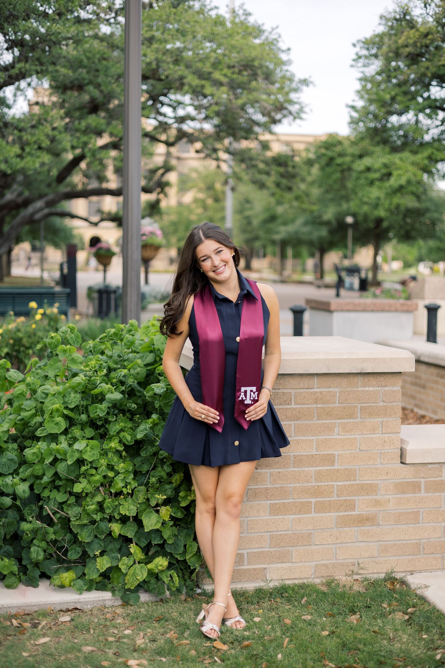 Texas A&M senior in navy dress and maroon graduation stole leans against brick wall among greenery.