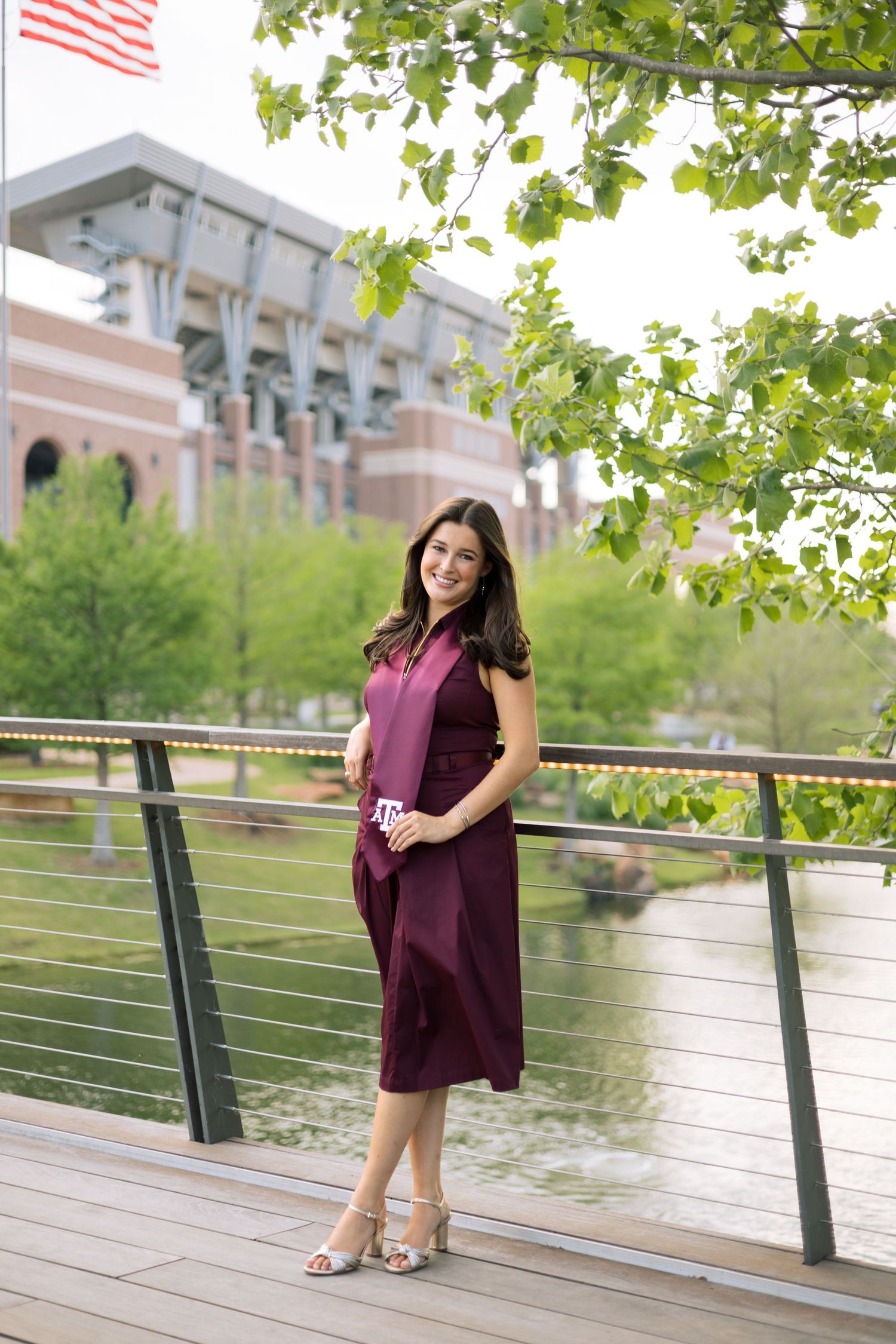 Texas A&M graduate in maroon dress posing on bridge with Kyle Field stadium in background.