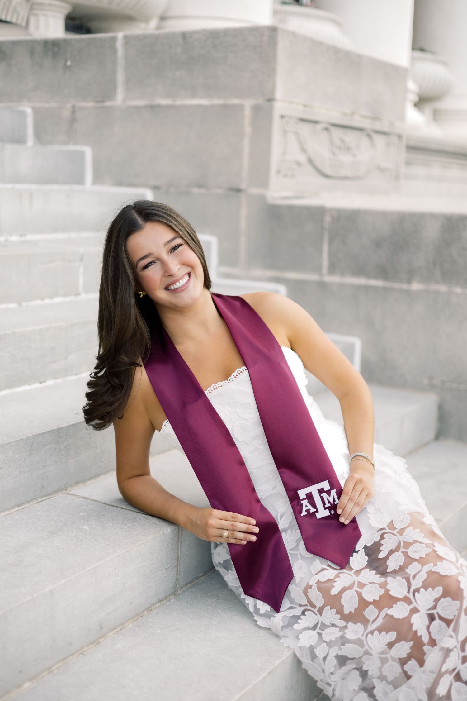 Happy graduate in white lace dress wearing Texas A&M maroon stole seated on stone steps.