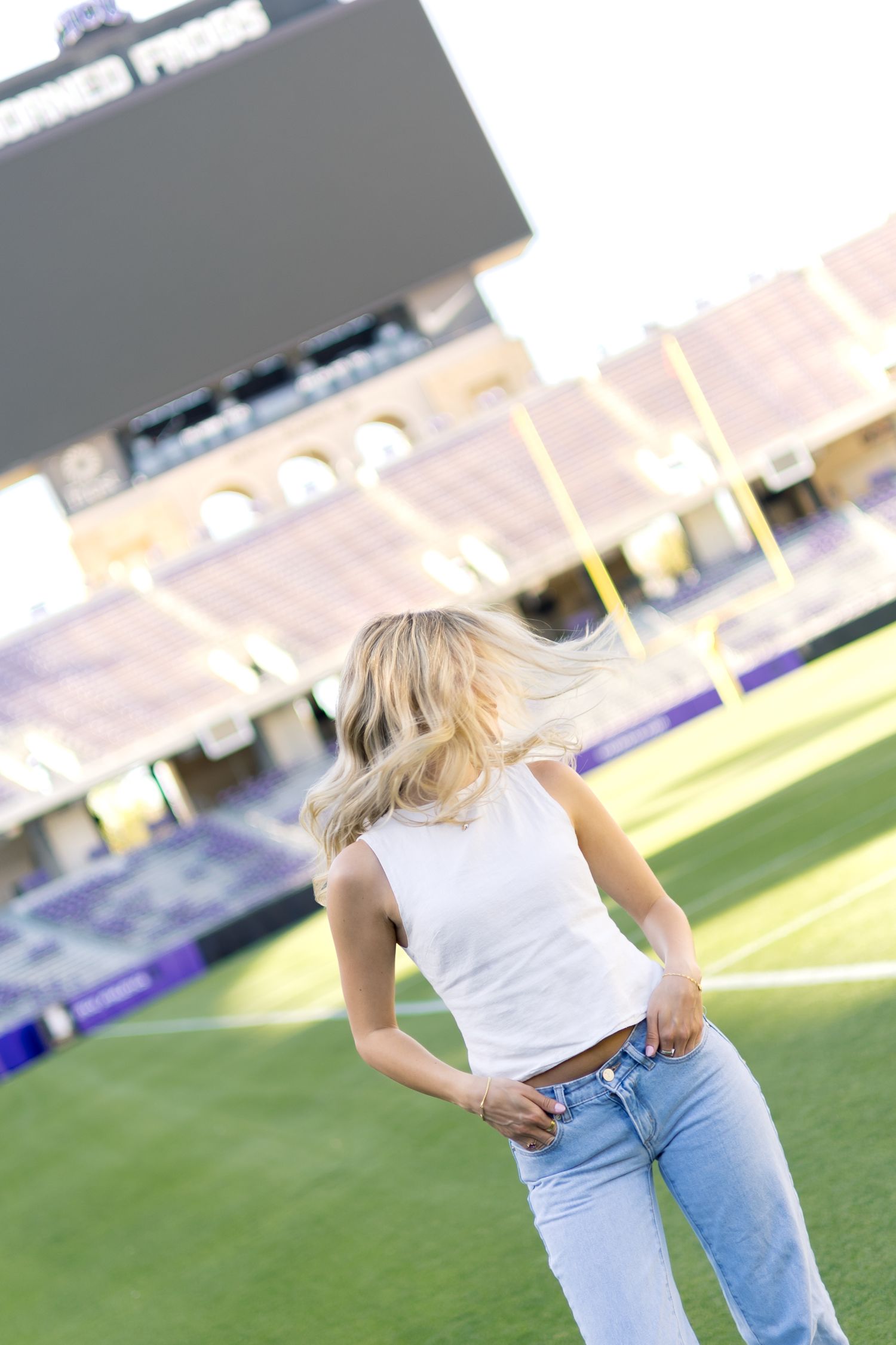 Blonde woman in white tank and jeans poses on green football field with purple stadium behind her.