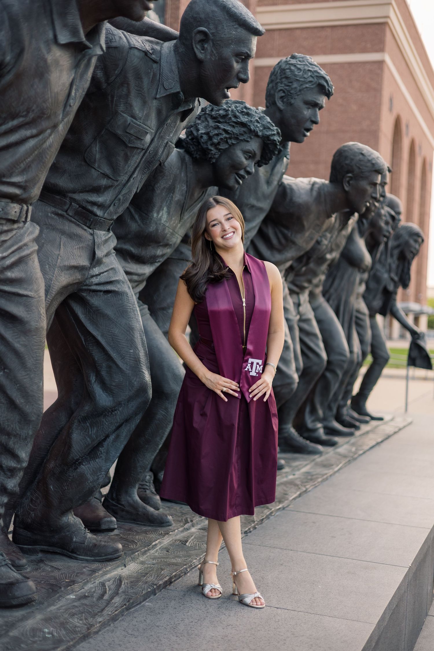 Texas A&M graduate in maroon dress stands proudly among bronze statues on campus.