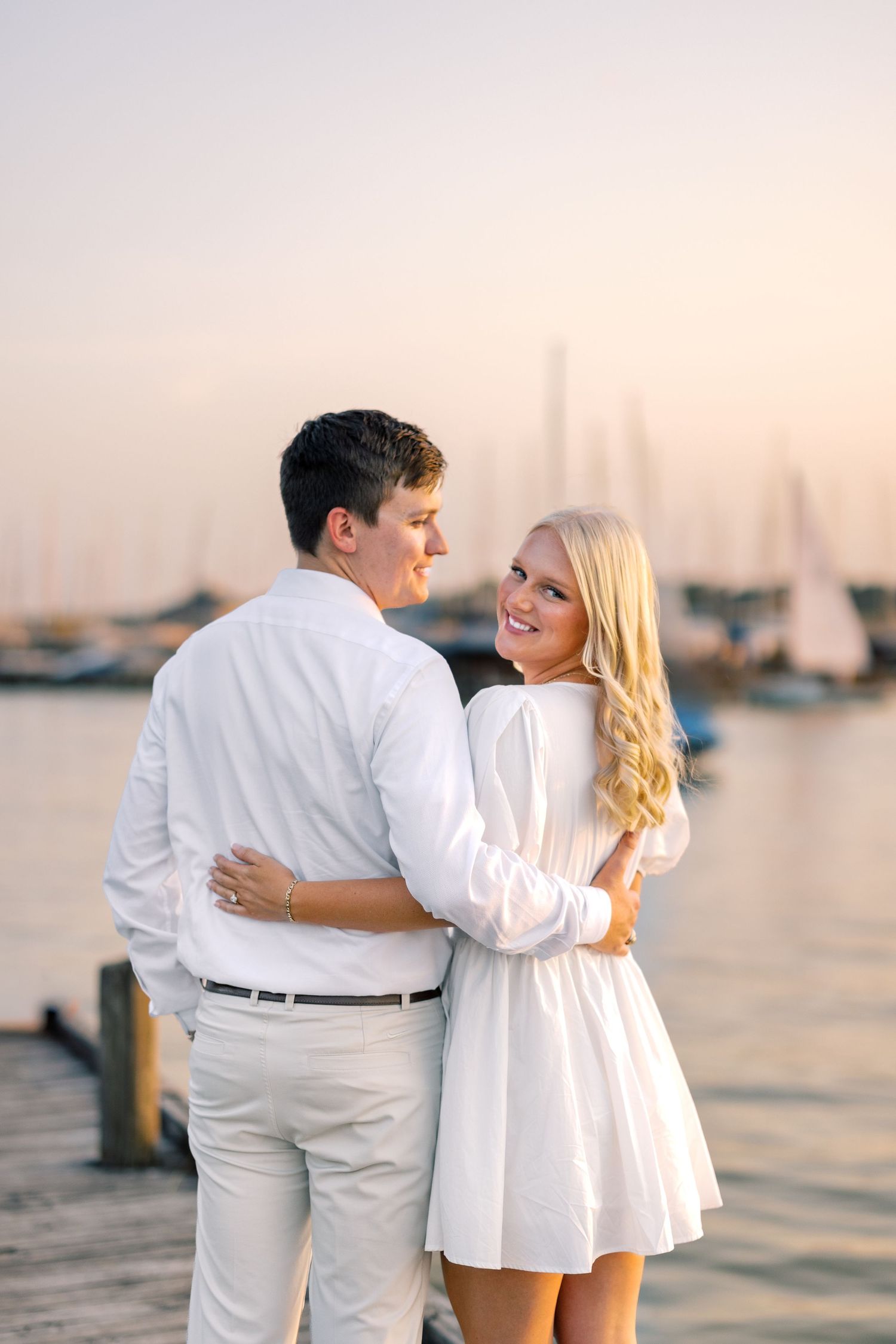 Couple in white outfits embracing on a marina dock at sunset with sailboats in the background.