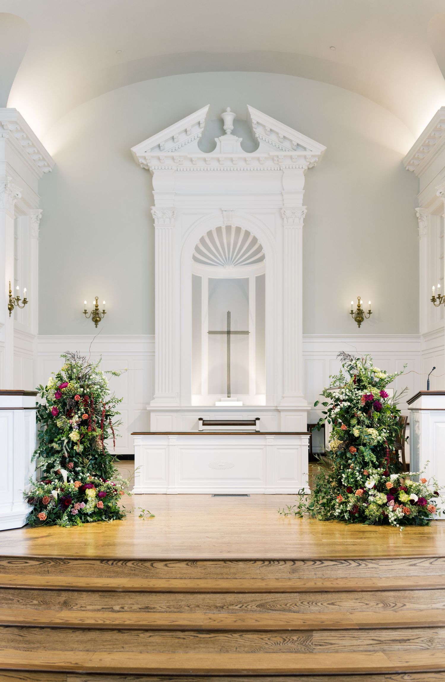 Elegant church altar adorned with lush floral arrangements flanking a white pediment with cross for a wedding ceremony.