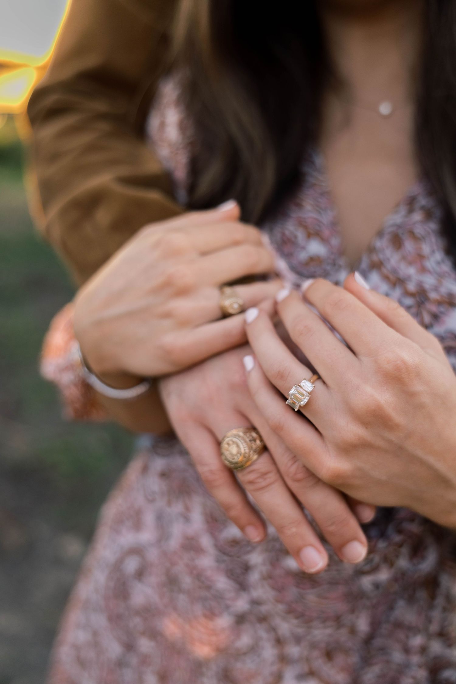 Close-up of couple's hands showing gold engagement ring and wedding band, woman in floral dress, man in brown jacket.