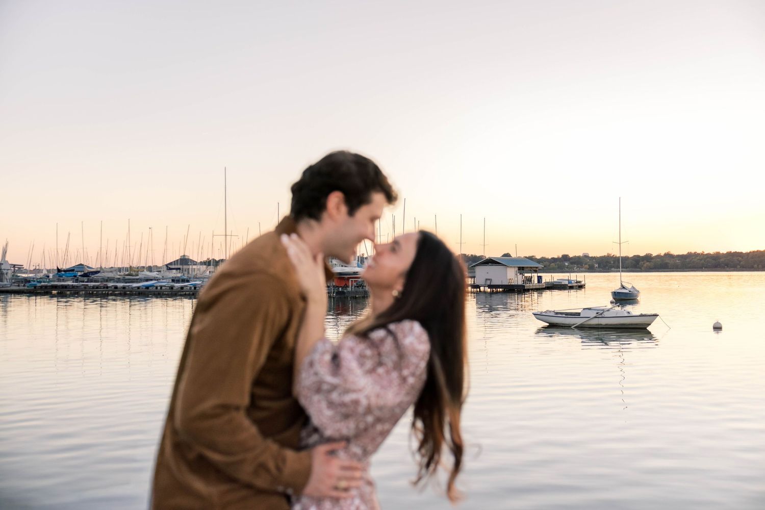 Romantic couple kissing at sunset by a serene harbor with sailboats anchored in the background.