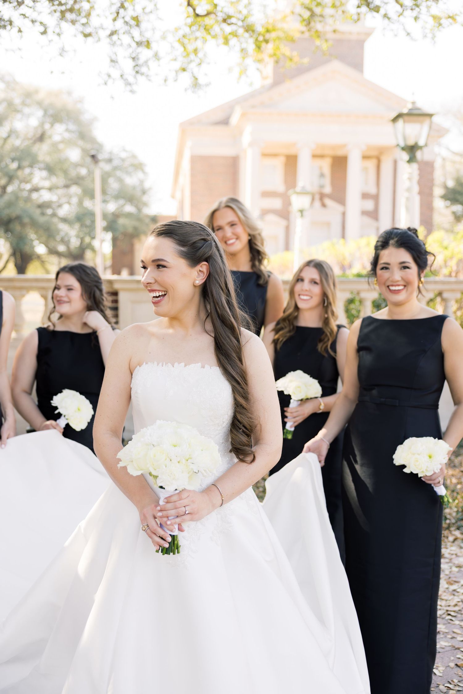 Bride in white gown laughing with bridesmaids in black dresses holding white bouquets.