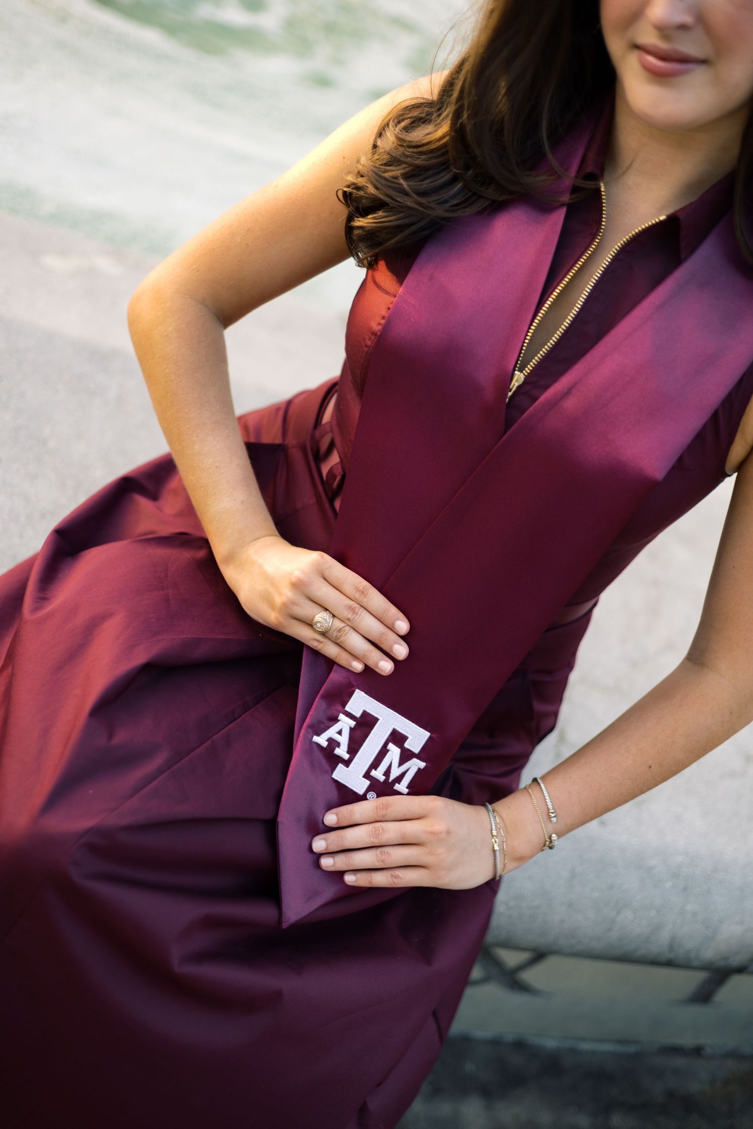 Woman in maroon Texas A&M graduation gown holding sash with ATM logo, wearing silver bracelet and ring.
