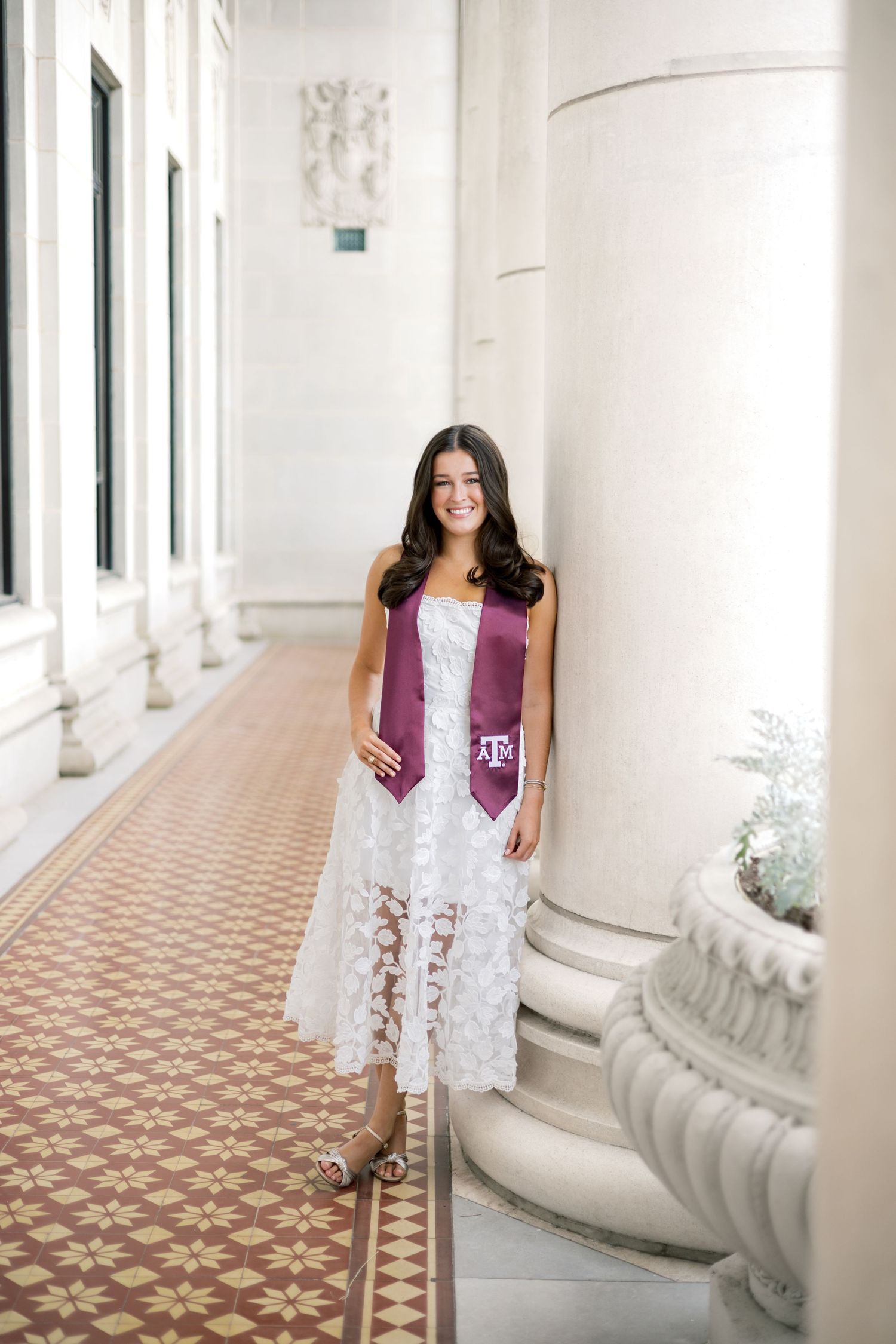 Texas A&M graduate in white lace dress with maroon ATM stole smiles in sunlit columned corridor.