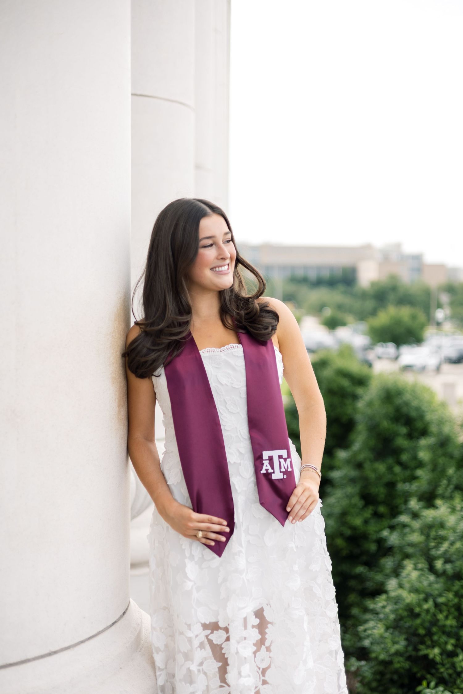 Brunette woman in white lace dress with Texas A&M maroon stole leaning against column with green trees in background.